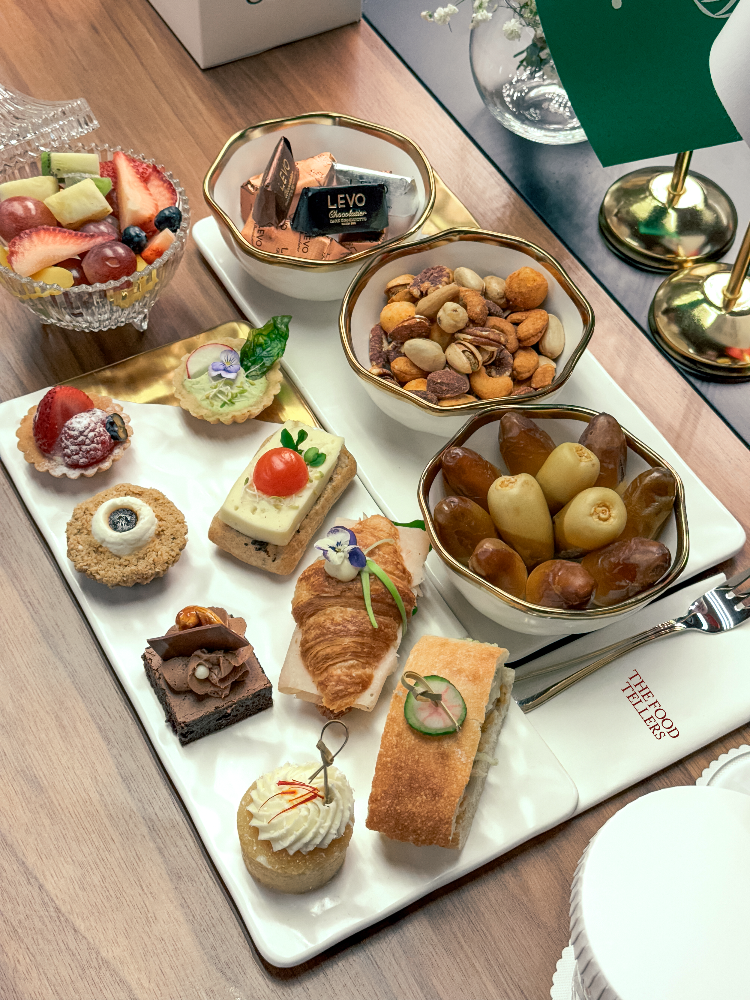 Assorted desserts and snacks on a white platter, including mini cakes, pastries, and candies, alongside bowls of mixed nuts, chocolates, and dried fruits on a wooden table.