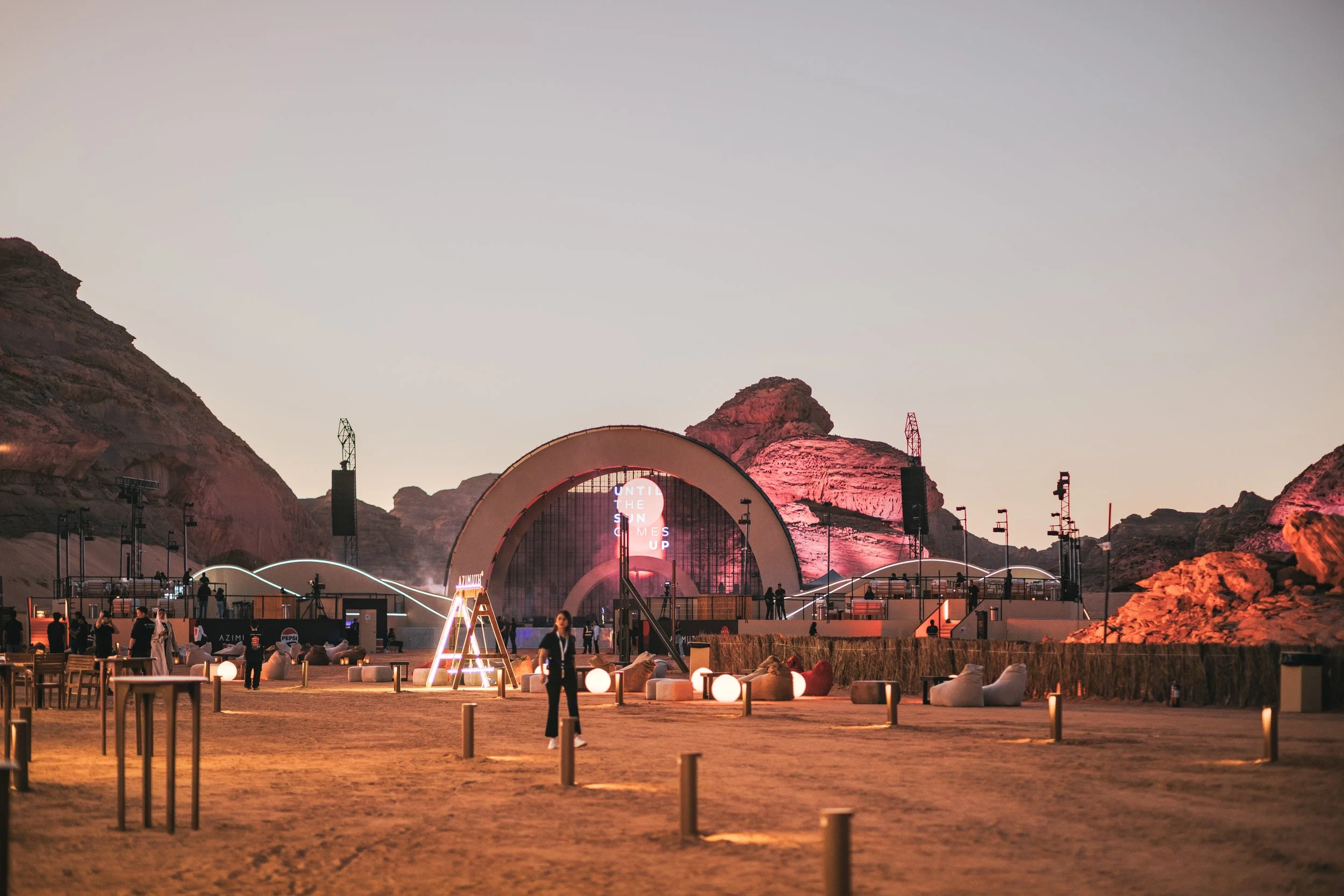 An outdoor concert or event venue set in a desert landscape with mountainous background during sunset or dusk. The stage features a large arch and colorful lighting, with scattered seating and people walking around.