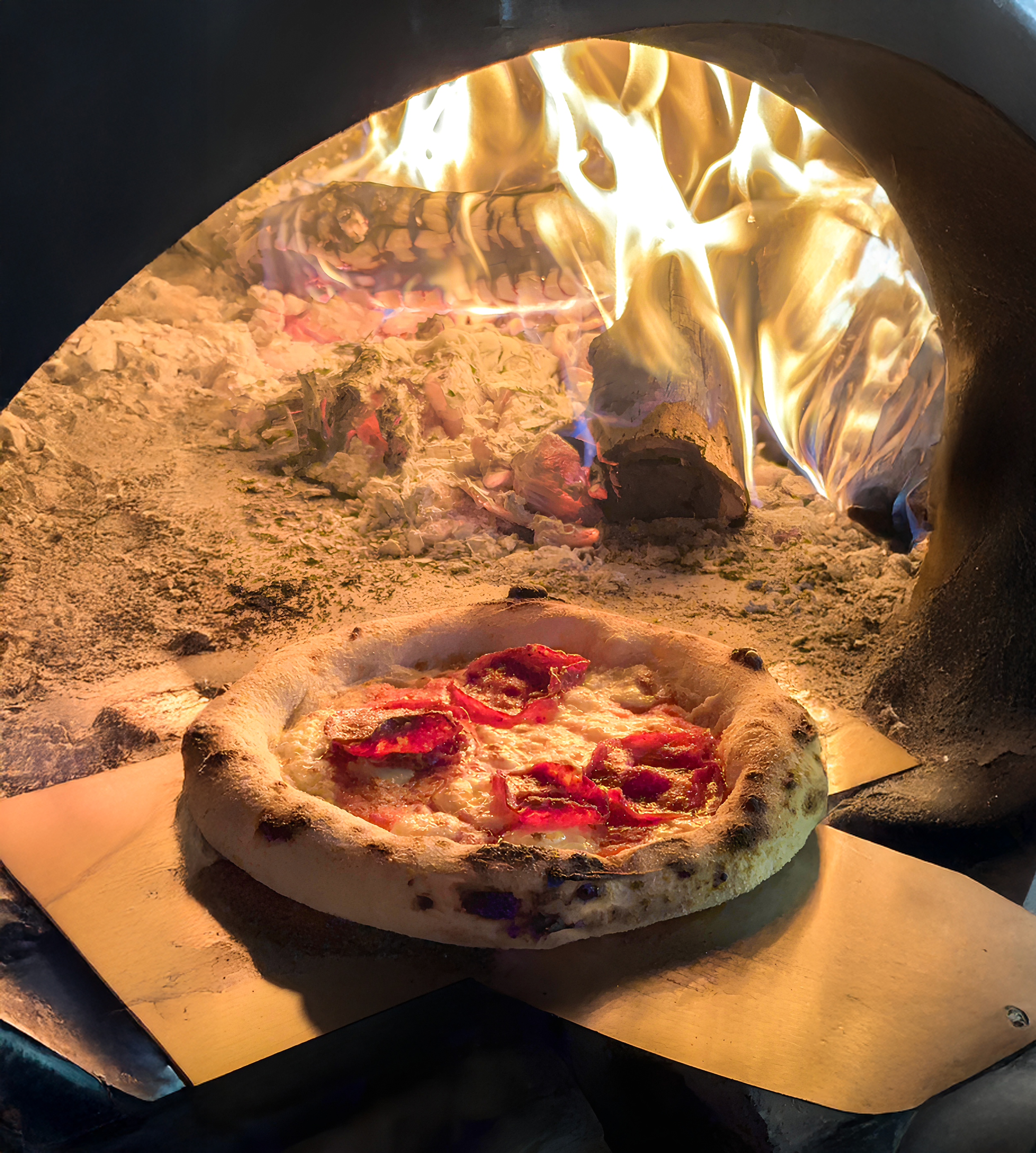 A pizza baking inside a wood-fired oven, with flames and ashes visible in the background.