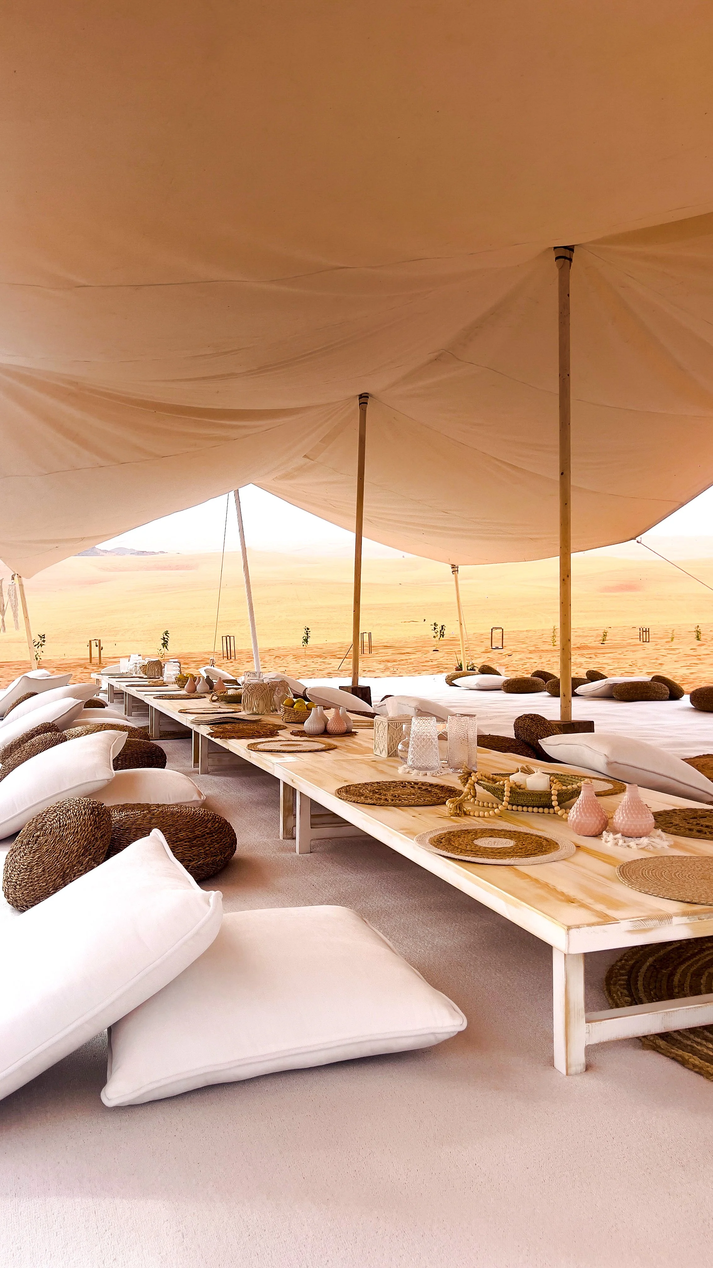 A desert tent setup with low tables, cushions, and decorative items, overlooking the sand dunes.