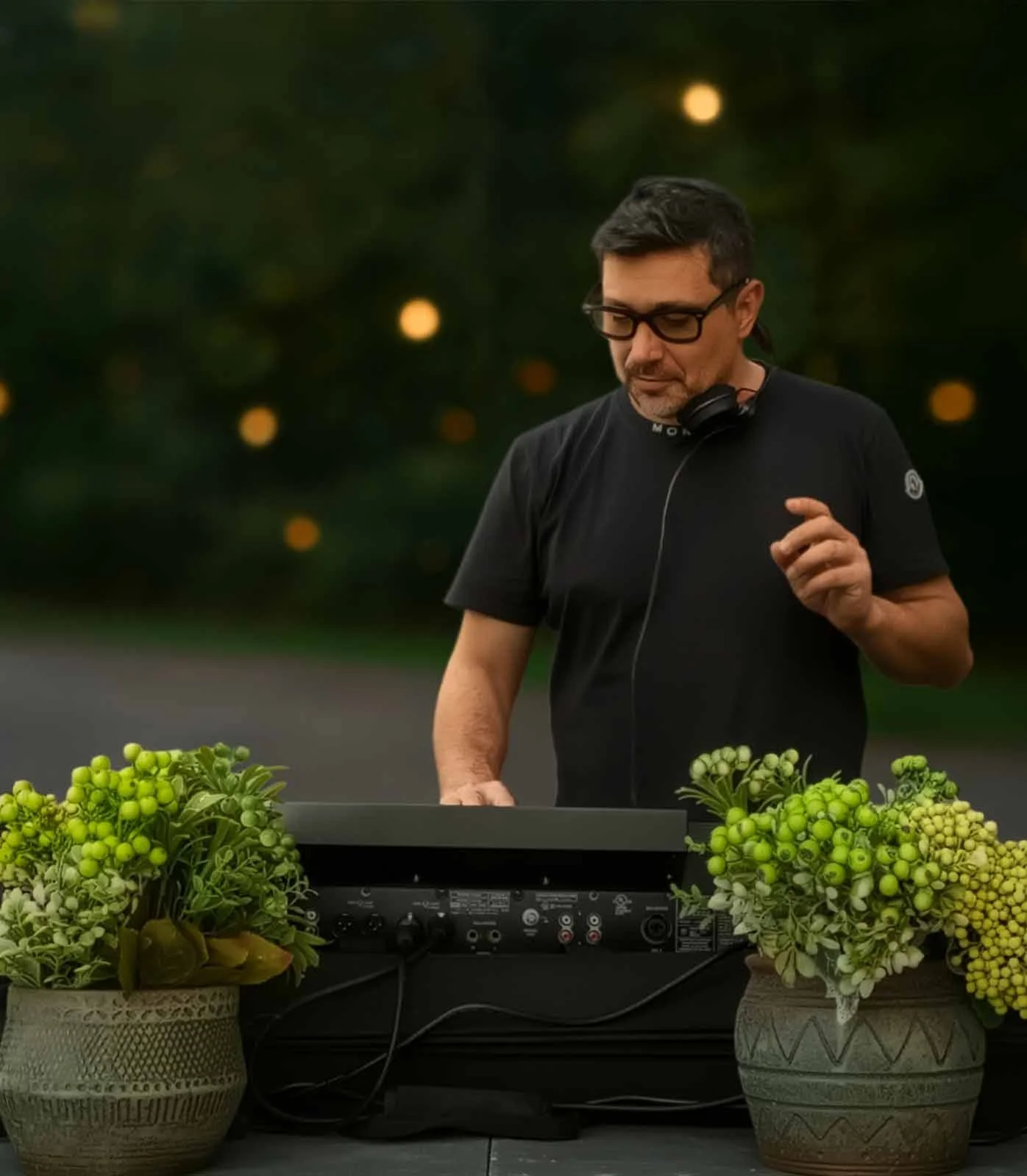 A man with glasses and headphones around his neck is DJing at an outdoor event in the evening. There are two potted plants with green flowers on either side of him.