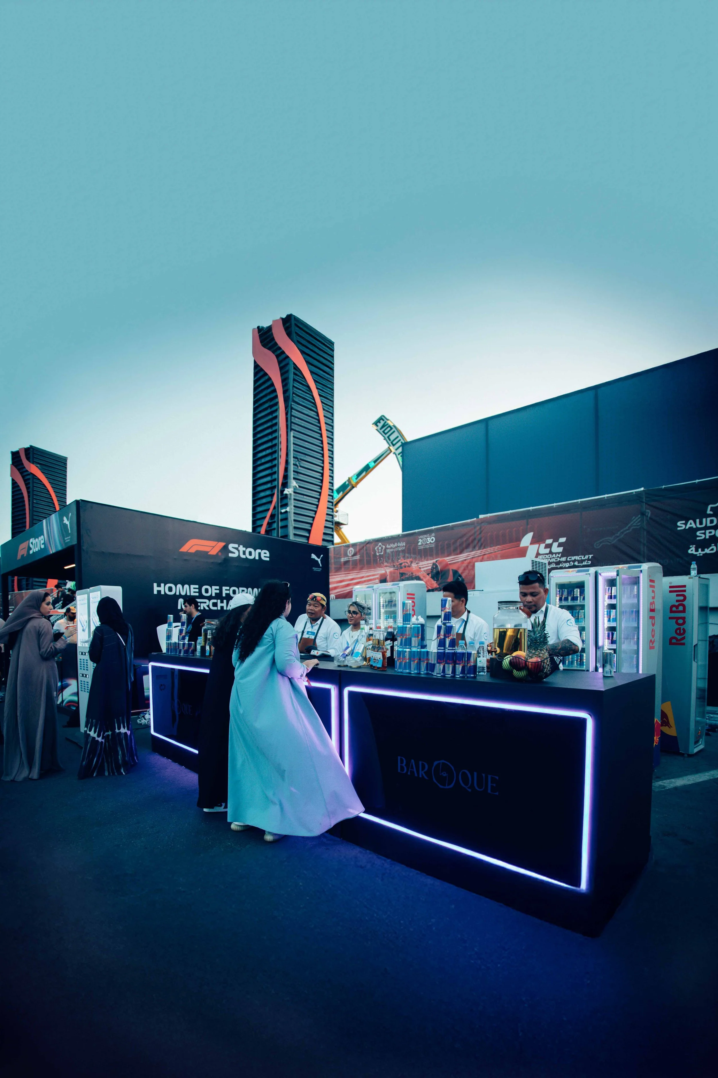 People at a Red Bull beverage stand at a Formula 1 event in an urban area with tall modern buildings and a clear sky.