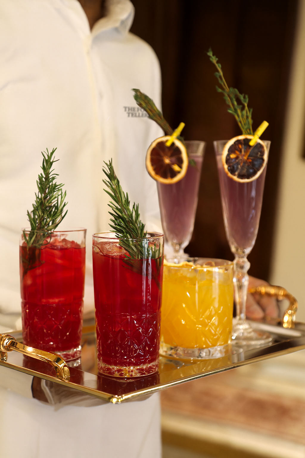 Assorted cocktails with garnishes on a tray, including red and yellow drinks with rosemary sprigs, and purple drinks with dried lemon slices and herbs.