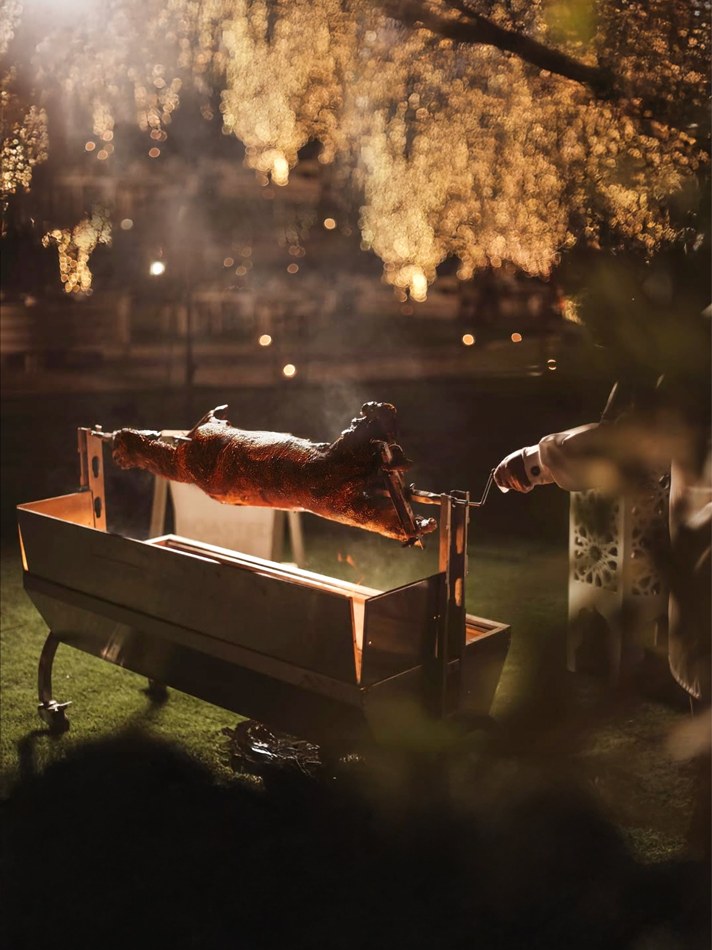 A roasted pig on a spit over a grill at night, illuminated by warm lighting, with trees and blurred lights in the background.