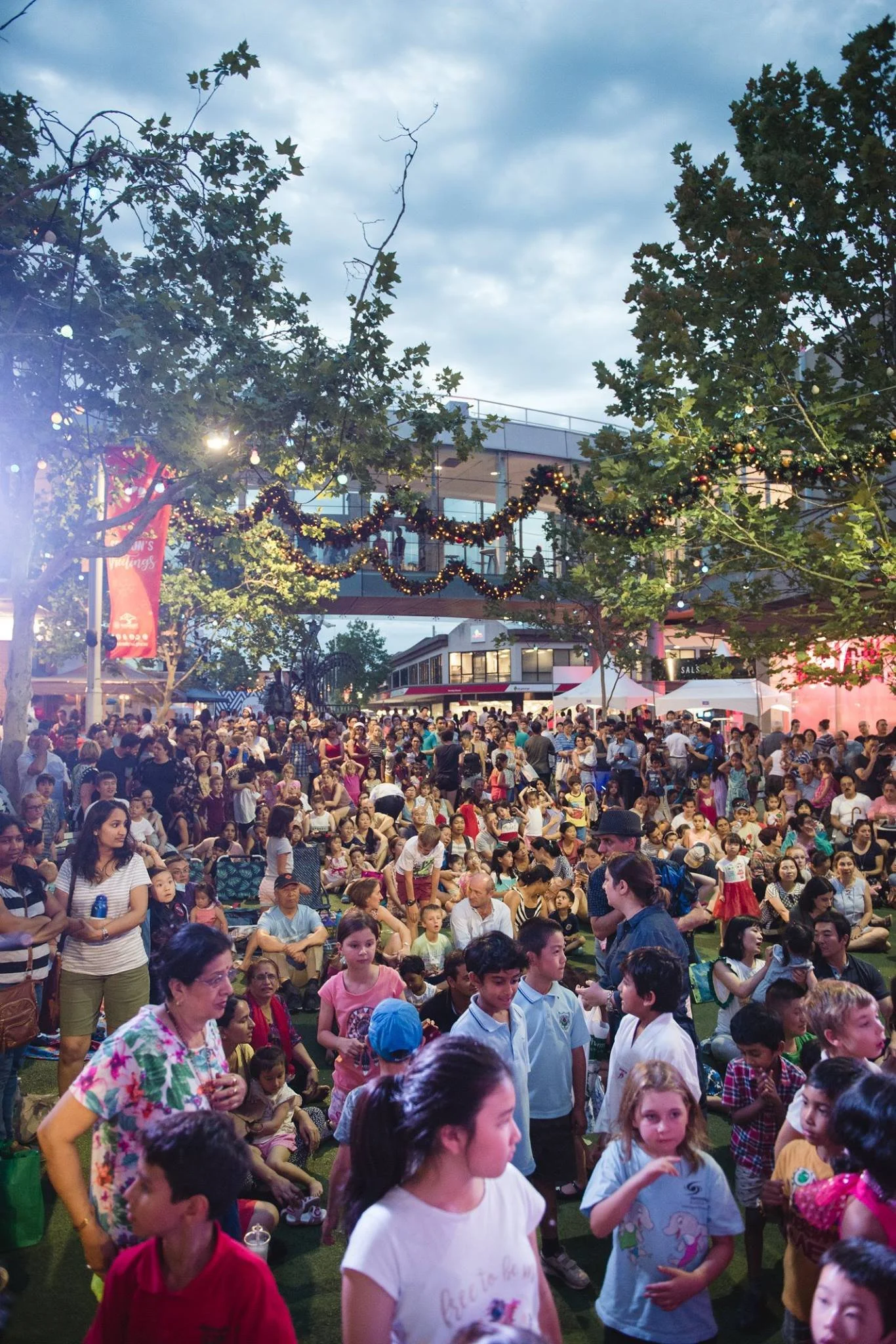Crowd of people attending an outdoor event with holiday decorations and string lights hung across trees and buildings, during the evening.