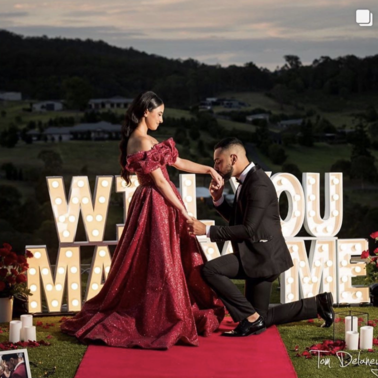 A man in a tuxedo proposes to a woman in a red gown outdoors, with large letter signs spelling 'WILL YOU MARRY ME' lit up behind them. The scene includes candles and flowers on a red carpet against a scenic landscape at dusk.