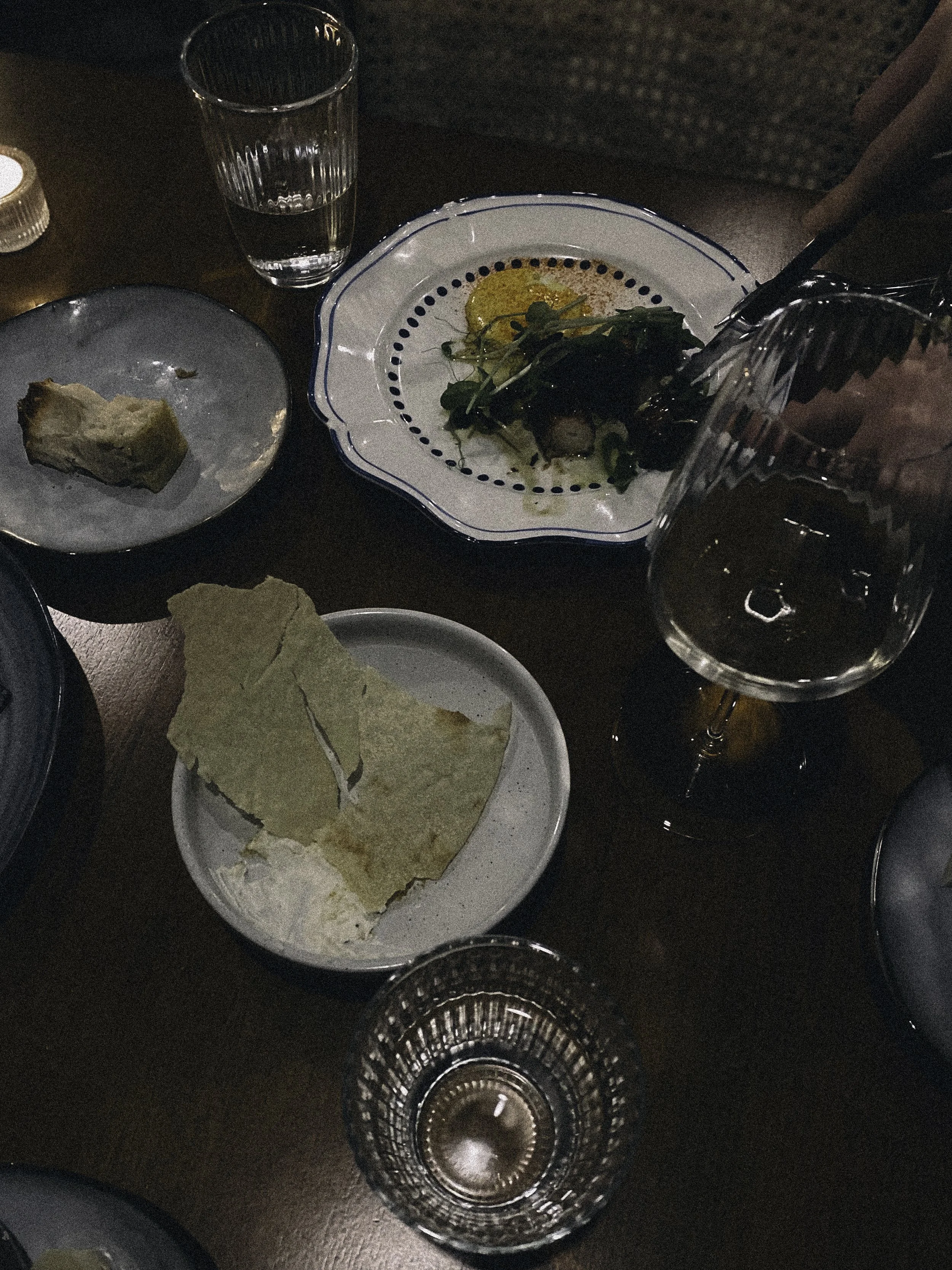 Dining table with plates of food, a glass of water, a wine glass, bread, and a small glass of what appears to be whiskey or similar liquor.