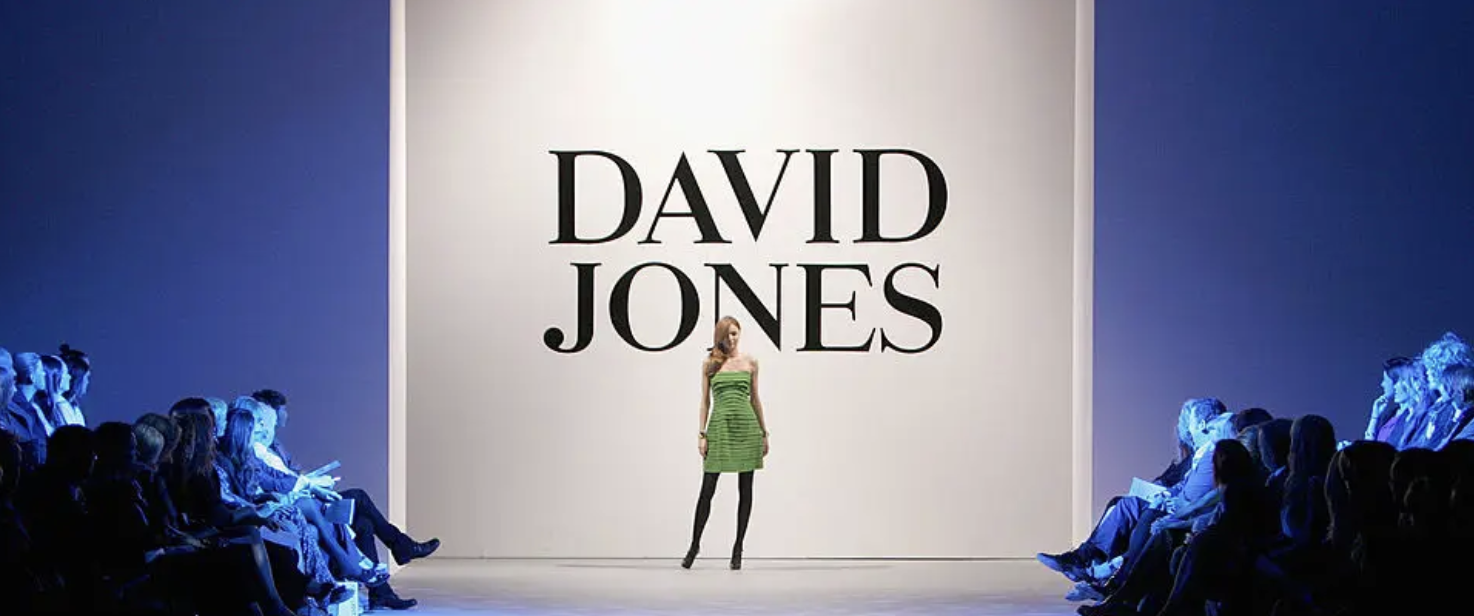 Fashion runway show with a model on stage in front of a large screen that reads 'David Jones,' with audience seated on both sides.