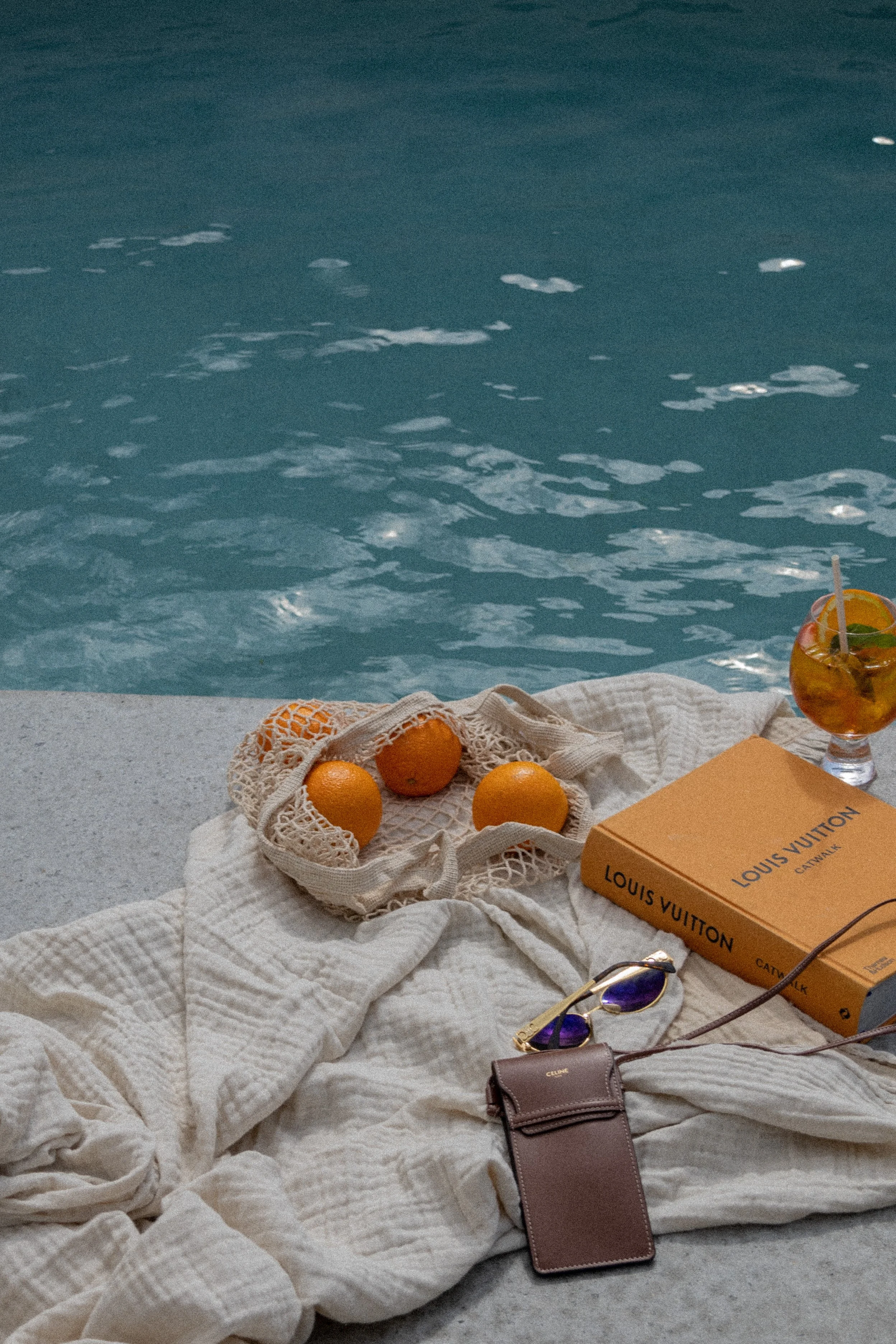 Poolside scene with a beige towel, oranges in a mesh bag, a Louis Vuitton book, sunglasses, a small brown pouch, and a cocktail with fruit on a concrete surface next to a swimming pool.