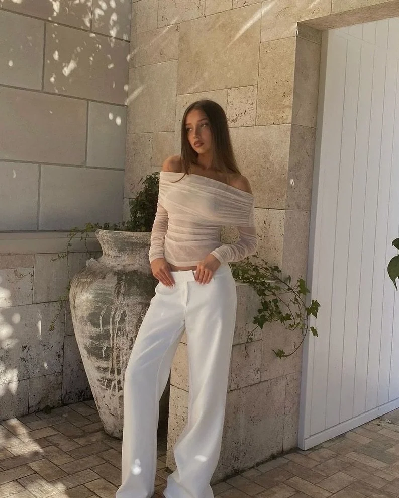 A young woman in a beige off-shoulder top and white wide-leg pants standing outdoors against a stone wall and white door, with sunlight and shadows.