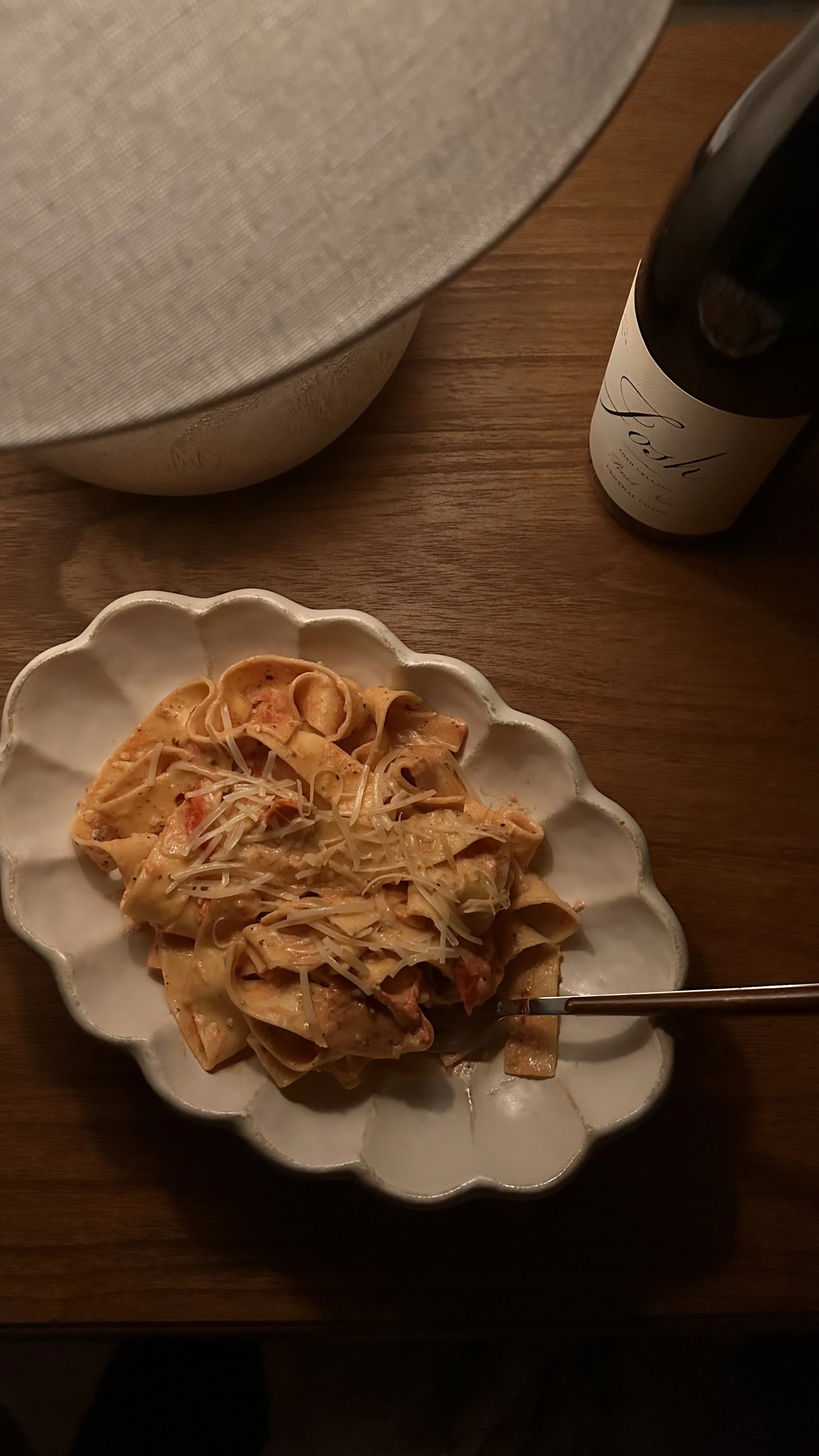 Plate of pasta with grated cheese and a fork, a bottle of wine, a container, and a bowl on a wooden table.