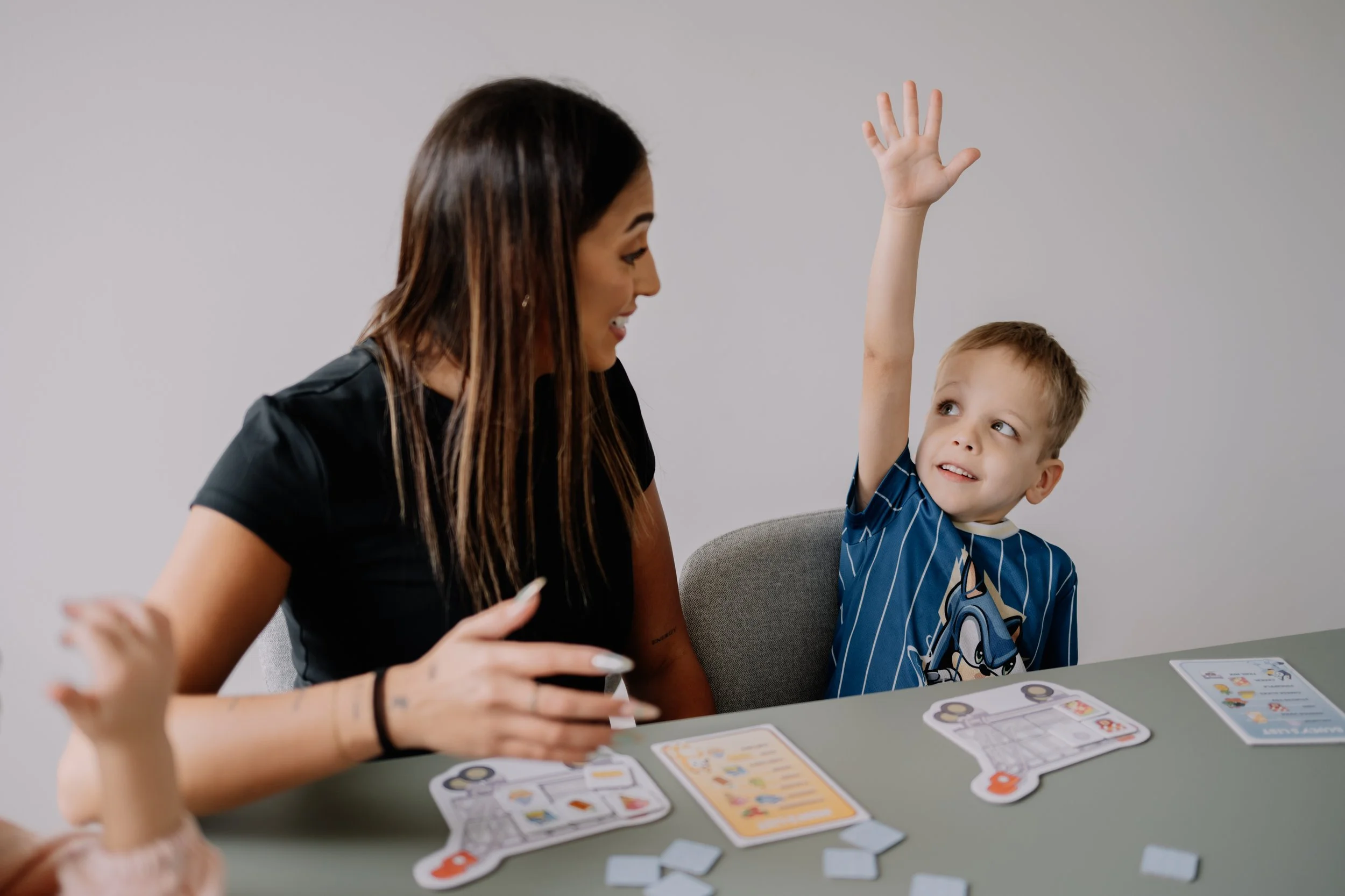 Eleyna and a young boy playing a board game at a table, with the boy raising his hand.