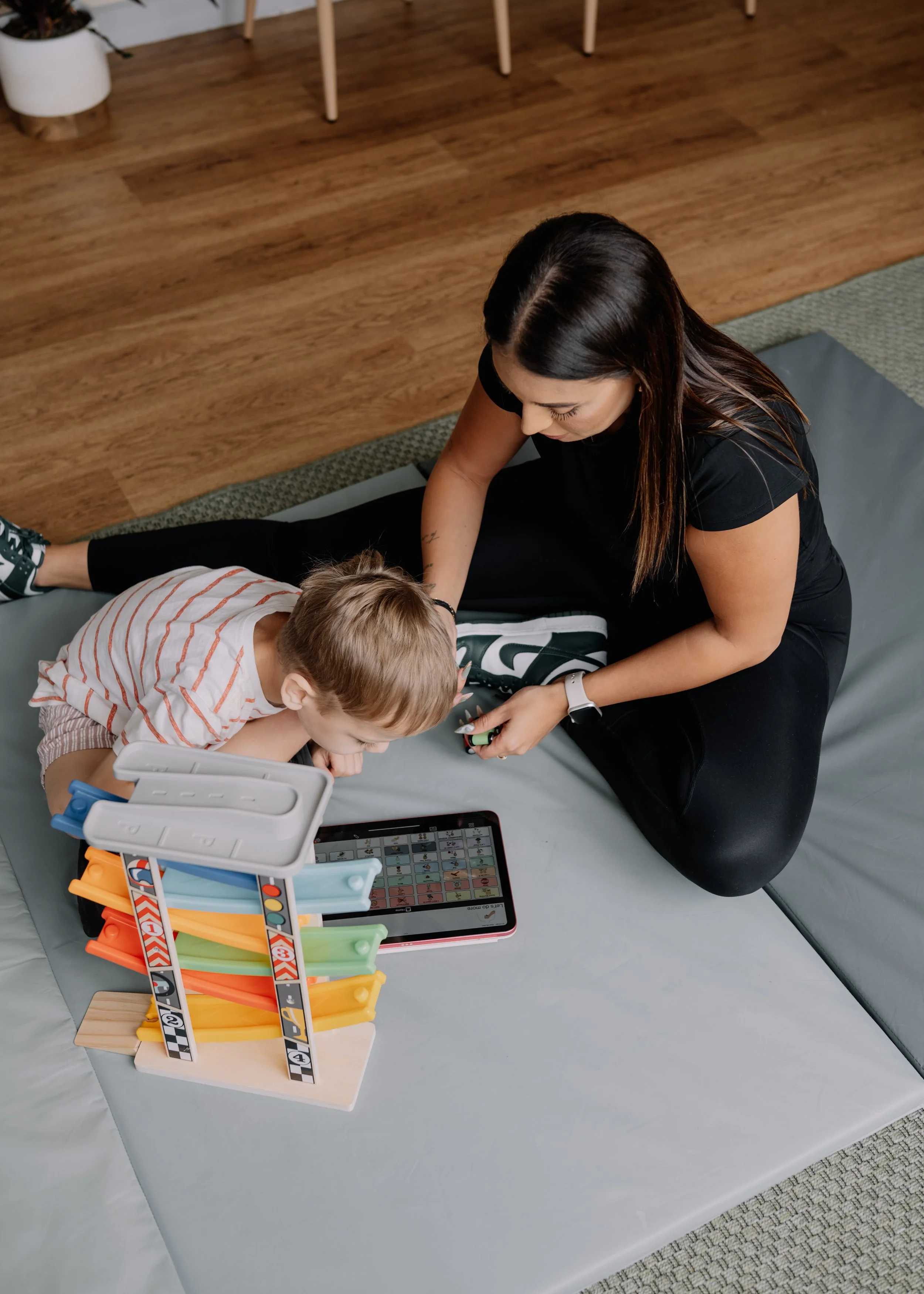 Eleyna and a young boy sitting on a padded mat, playing with a tablet and a toy racing track, in a room with wooden flooring.
