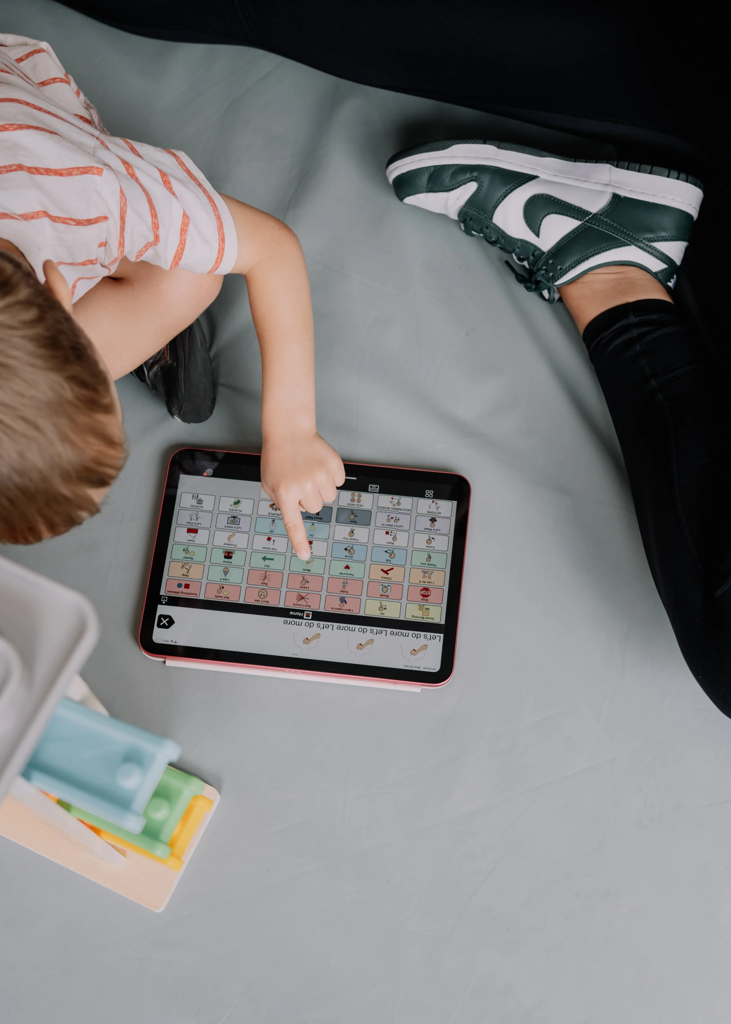 A young child with light brown hair and a striped shirt interacts with an electronic communication device, sitting on a gray surface, with an adult wearing black pants and sneakers nearby.