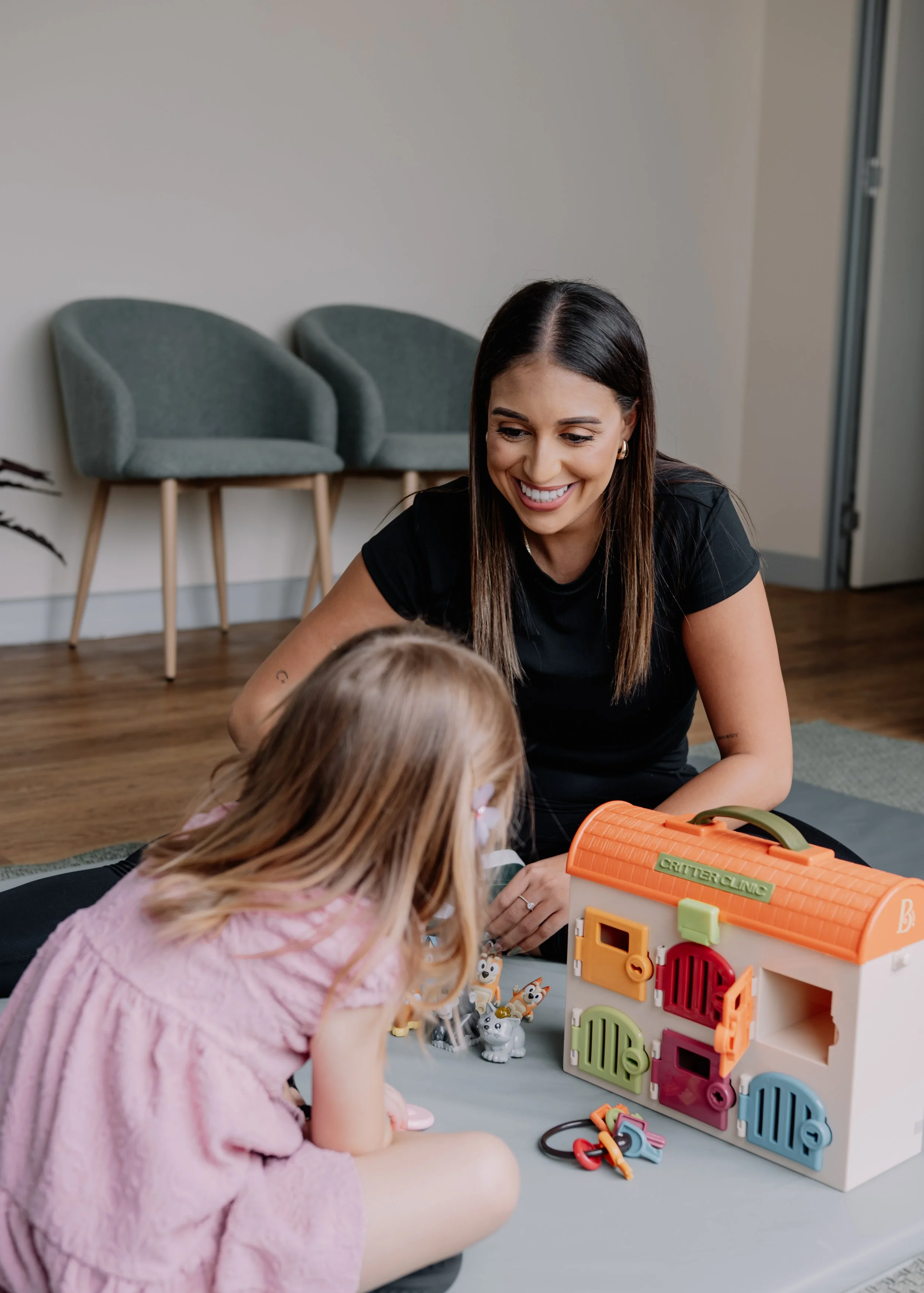Eleyna from Eleven Speech Pathology is playing with a young girl on the floor with a colorful toy house and animal figurines.