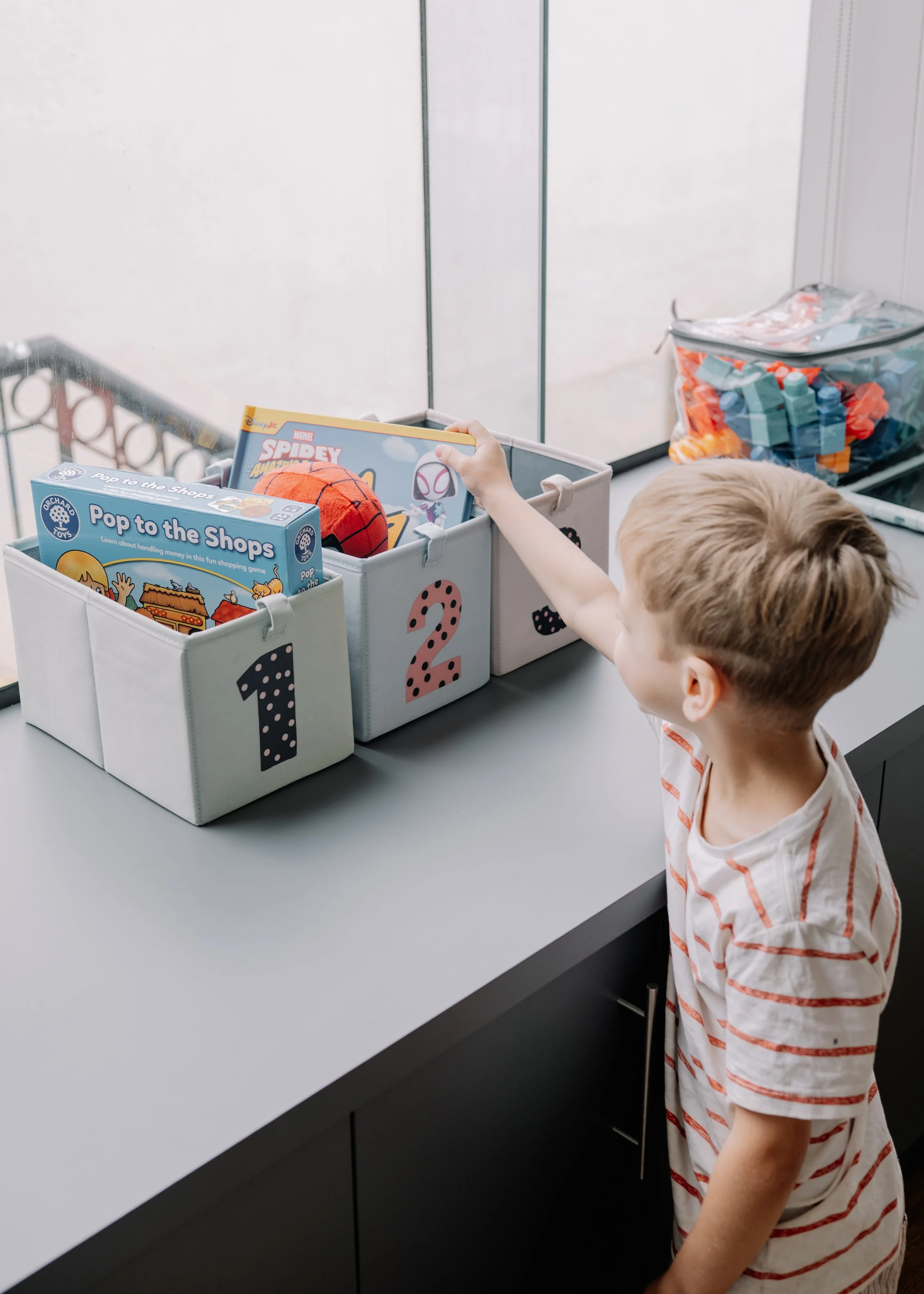 A young boy with blond hair wearing a white and red striped shirt reaching into a storage bin labeled '2' with a pink polka-dotted number. The bin contains children's books and a ball. There are similar bins labeled '1' and another with lego blocks i