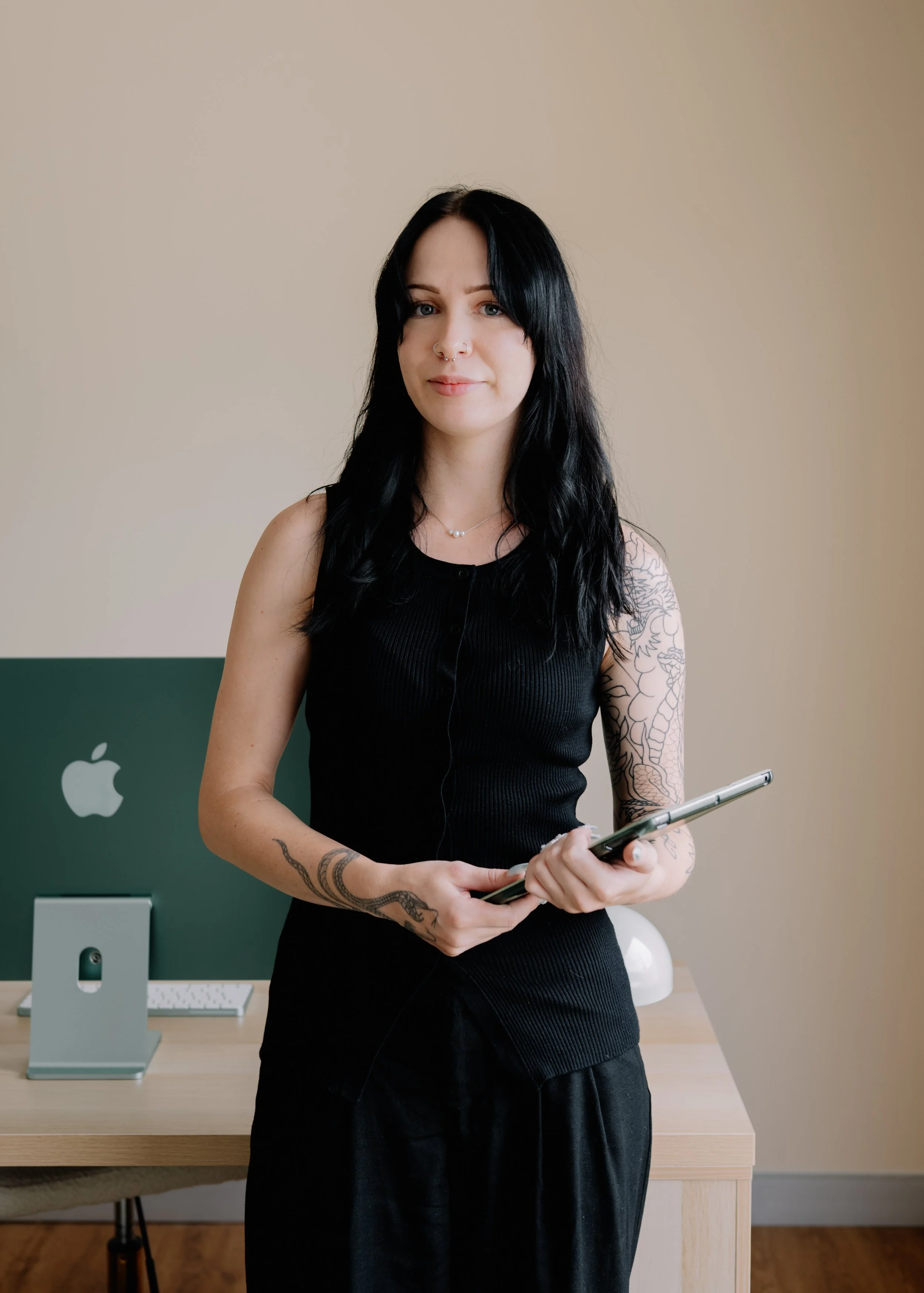 A woman with black hair, tattoos on her arms, wearing a black sleeveless top, holding a tablet, standing in front of a desk with Apple equipment and a beige wall.