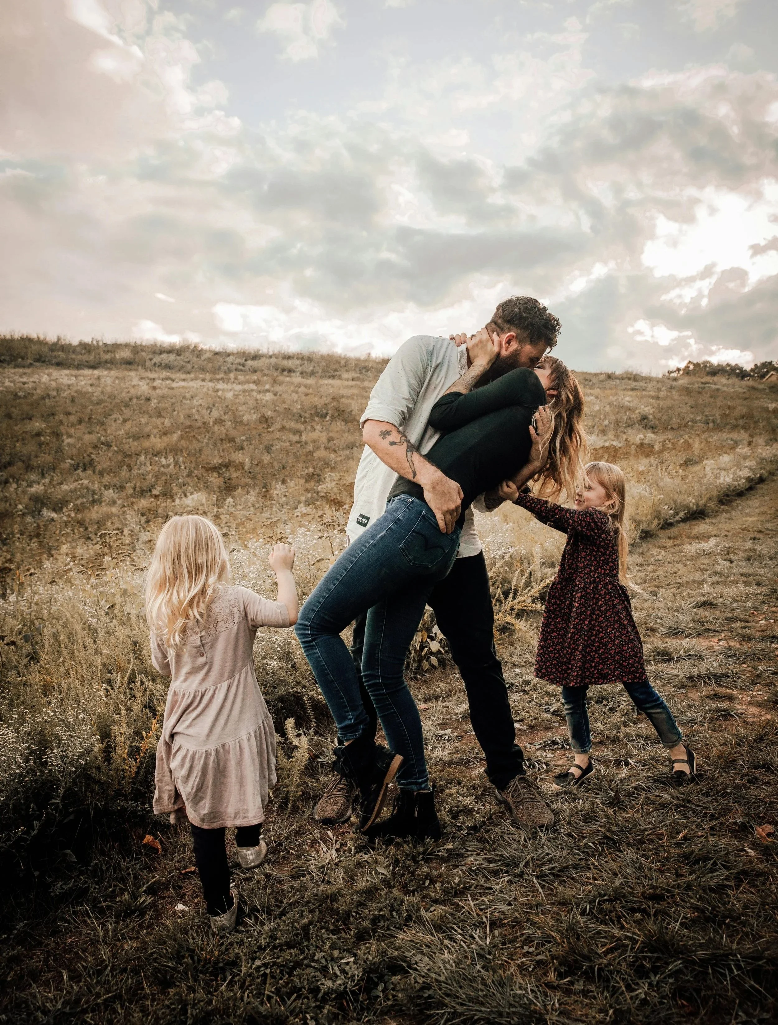 A couple kissing in an open field with two young girls nearby, one reaching out and the other walking away.