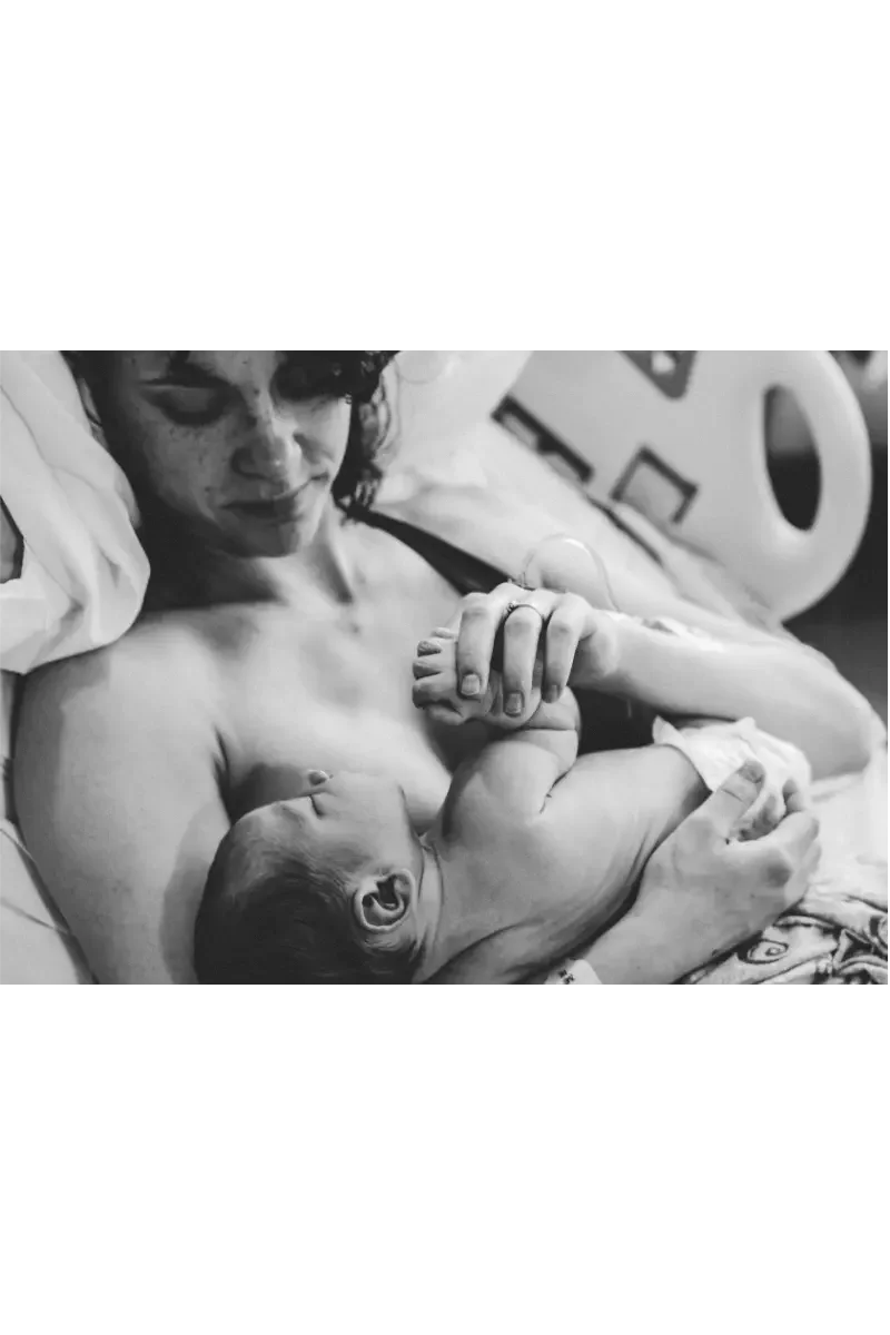 Black and white photo of a woman lying in a hospital bed, holding a newborn baby close to her chest, looking at baby.