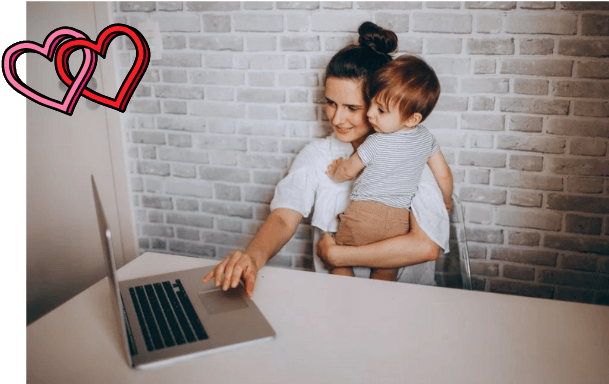 Woman sitting at a white table using a laptop, holding a young child in her lap who is hugging her head, with a brick wall in the background and two digital heart icons in the top left corner.