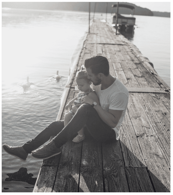 A dad and a young daughter sitting on a wooden dock by the water, with the dad holding the daughter close as they look at the water, and boats and ducks visible in the background.