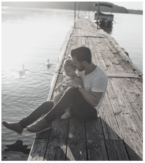 A dad and a young daughter sitting on a wooden dock by the water, with the dad holding the daughter close as they look at the water, and boats and ducks visible in the background.