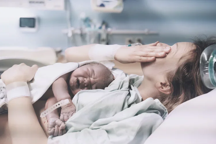 A brand new mom post-delivery, lying in a hospital bed, with a newborn baby on her chest, emotionally wiping her tears after giving birth.