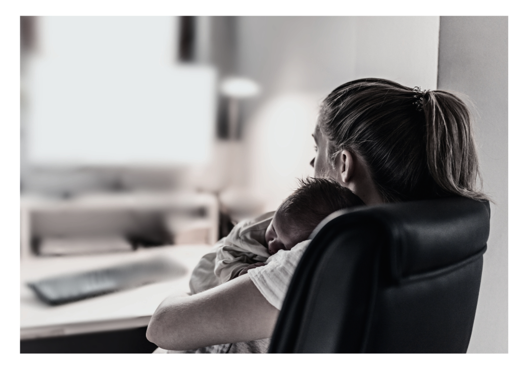 A new mom sitting in an office chair in front of a computer, holding and comforting a sleeping newborn, indicating doing online therapy.