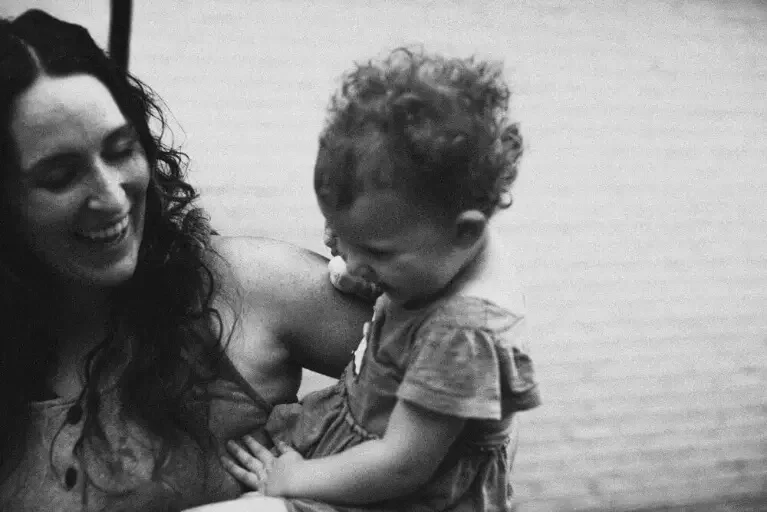 A smiling and laughing mom and toddler-aged baby on her hip, playing with a young child in a relaxed setting.