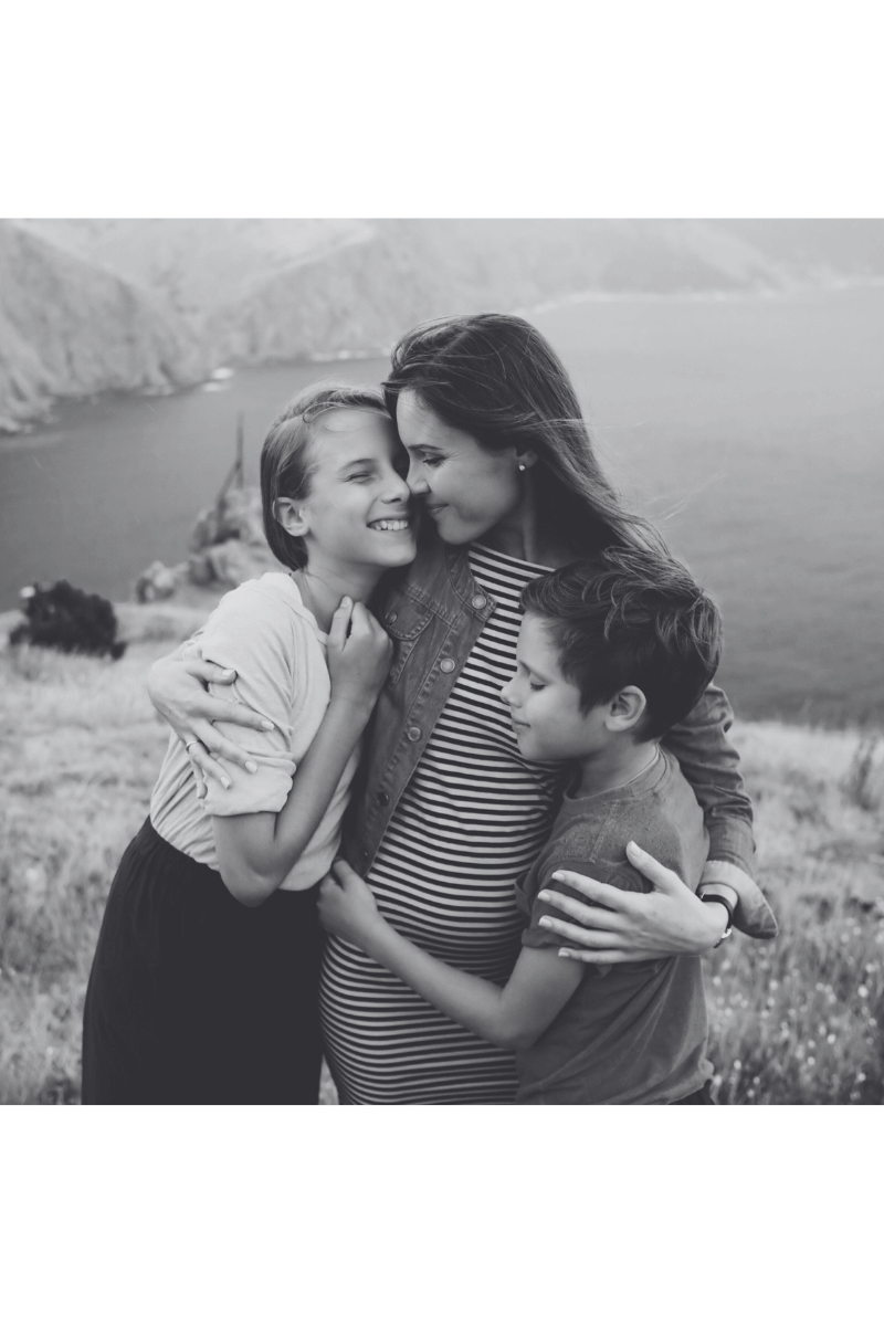 A pregnant mom with two older children, a girl and a boy, hugging on a grassy hilltop with a scenic view of a body of water and cliffs in the background.