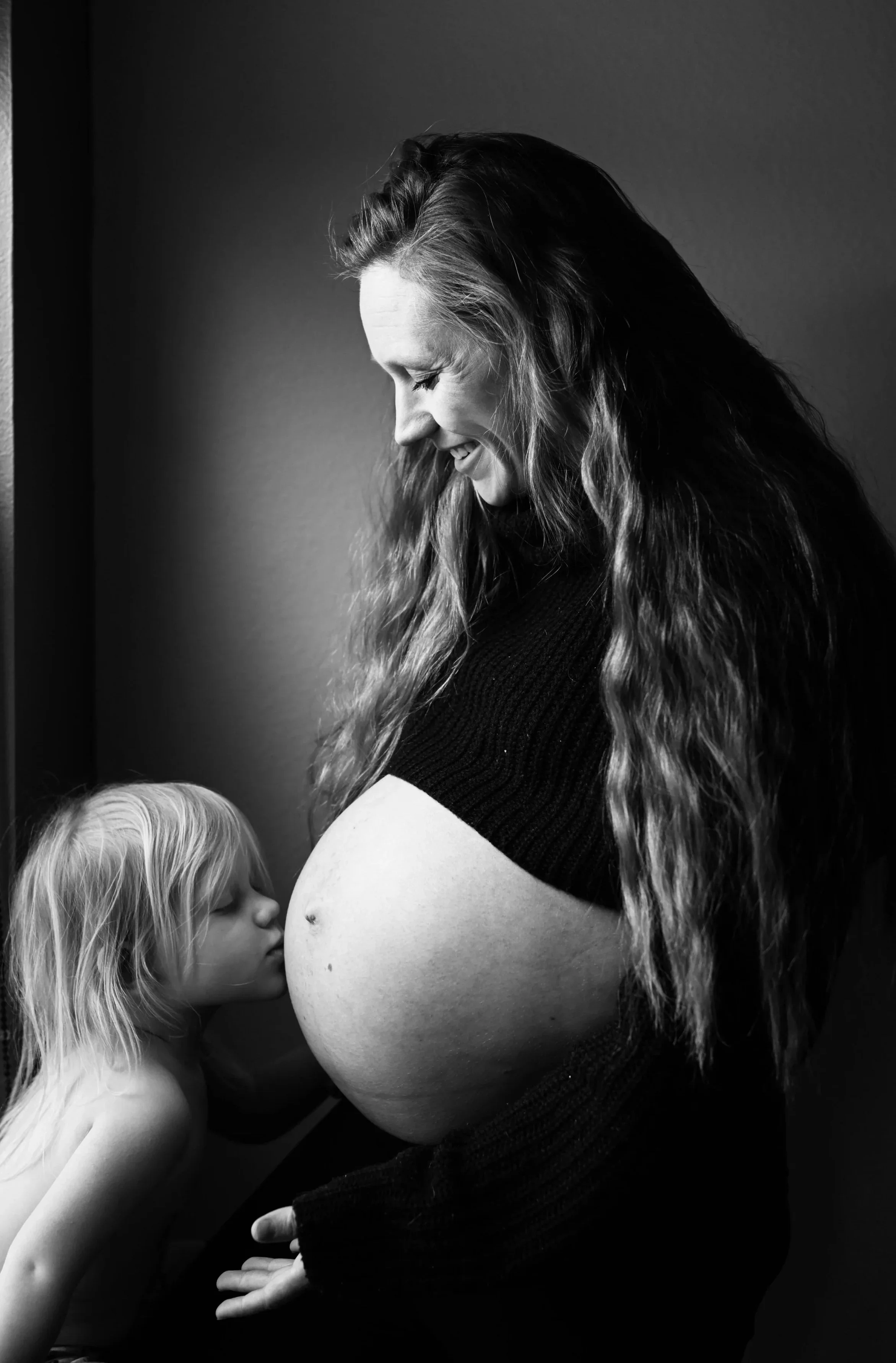 A pregnant woman with long wavy hair smiling and looking down at her small son, who is kissing her belly.
