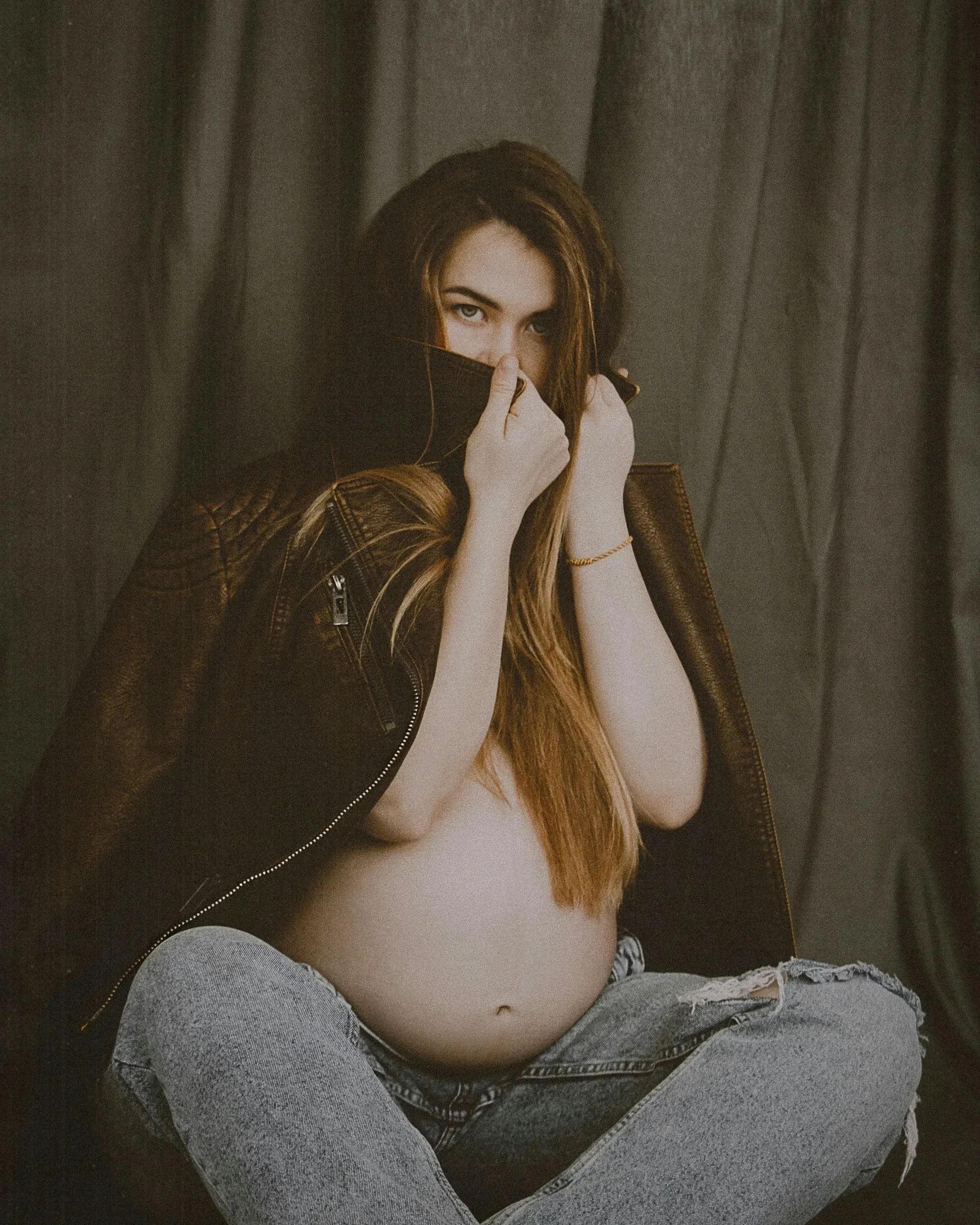 A pregnant woman with long brown hair sitting against a dark curtain, wearing a leather jacket and ripped jeans, partially covering her face with the jacket's collar and looking confident.