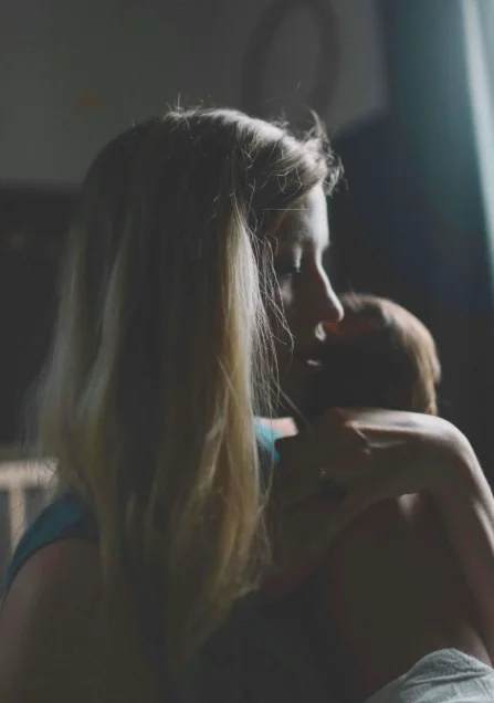 A new mom holds a small infant close to her face in a dimly lit nursery, looking on, indicating mixed emotions in postpartum.