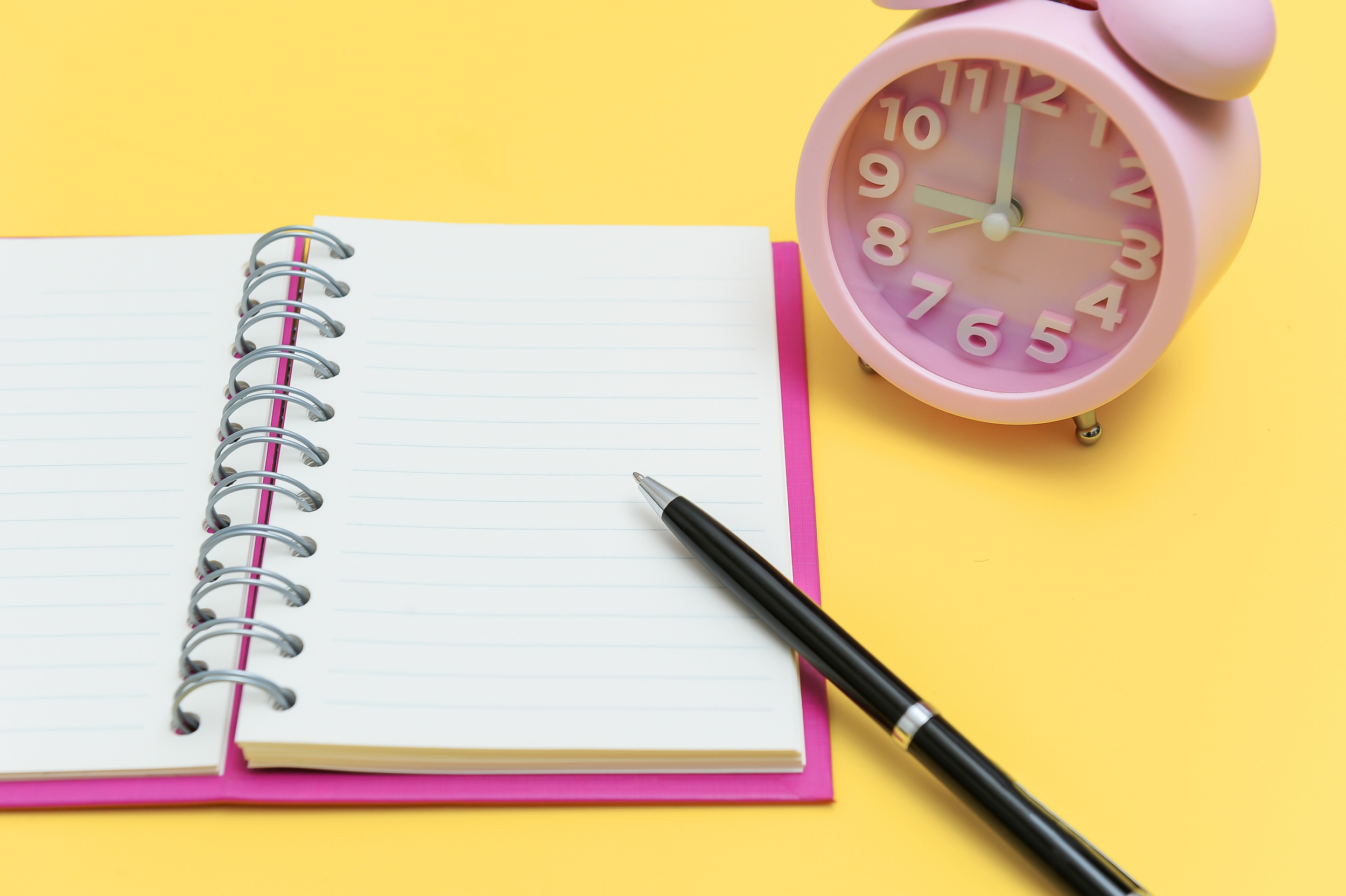 Open spiral notebook with blank lined pages, black pen resting on it, pink alarm clock showing 12:00, on a yellow background.