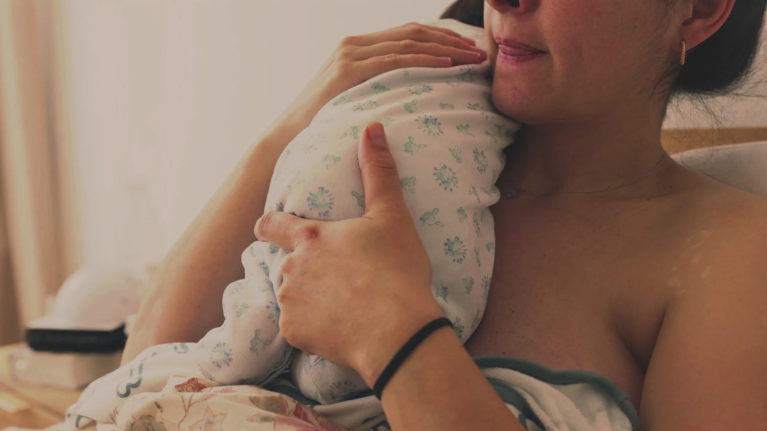 A woman holding a newborn baby in a hospital or home setting. The woman appears to be gently cradling the baby against her shoulder.