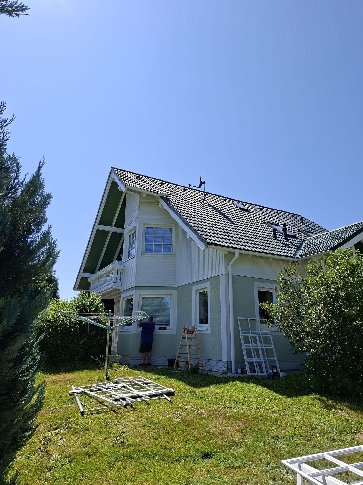 Person steht vor dem Fenster eines Hauses und arbeitet. Es sind Bau- oder Renovierungsarbeiten im Gange, mit zusammengelegtem Gerüst und Bauzubehör auf dem Rasen.