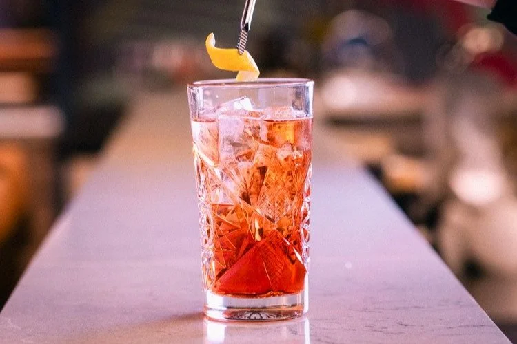 A tall glass filled with a pink-colored beverage, ice cubes, and a lemon wedge garnish, with a striped straw.