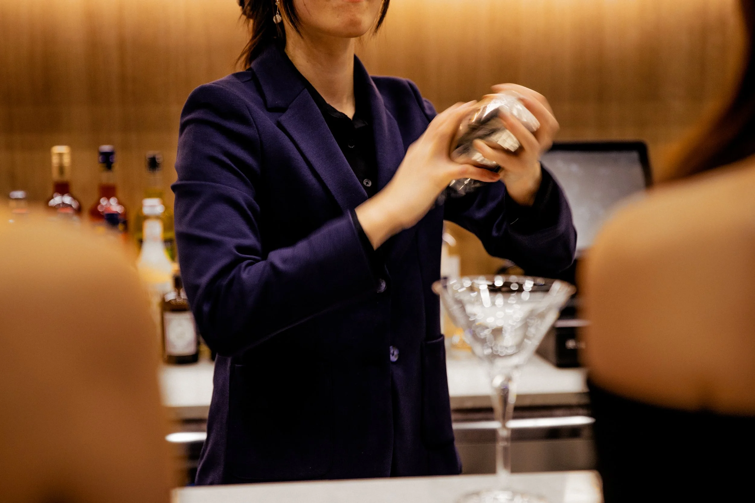 A person in a dark blazer preparing a cocktail at a bar, with bottles of alcohol in the background and a martini glass in the foreground.
