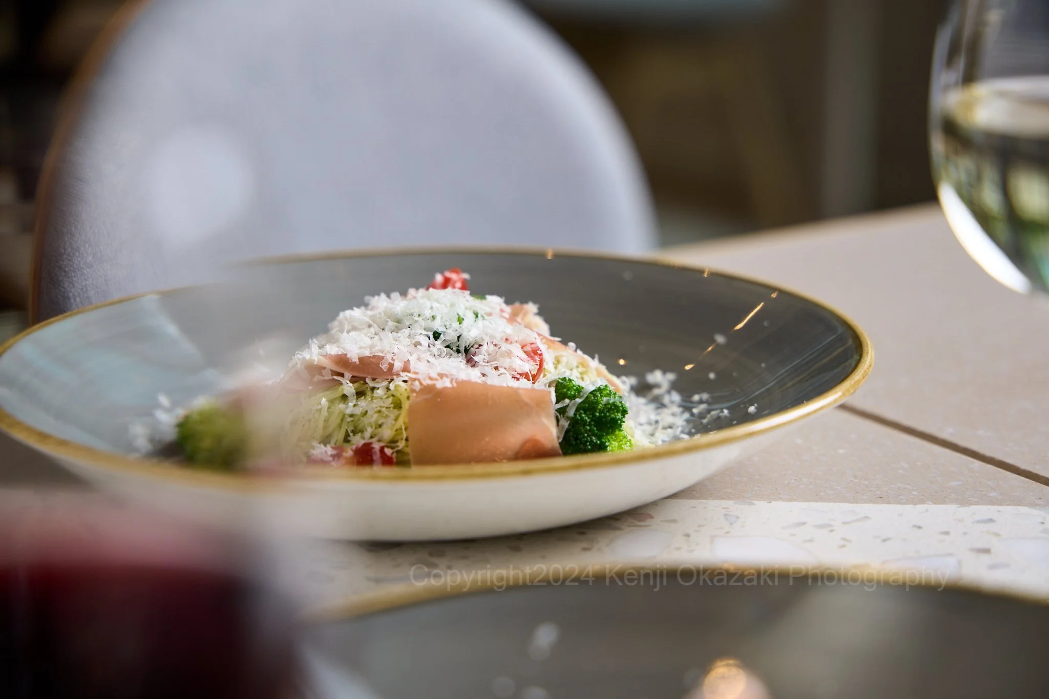 A plate of salad with shredded cheese, broccoli, ham, and tomatoes on a beige table with a glass of white wine in the background.