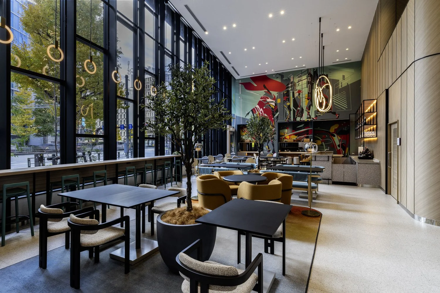 Modern restaurant interior with large windows, trees outside, colorful abstract wall art, trees inside planters, various tables and chairs, and contemporary lighting fixtures.