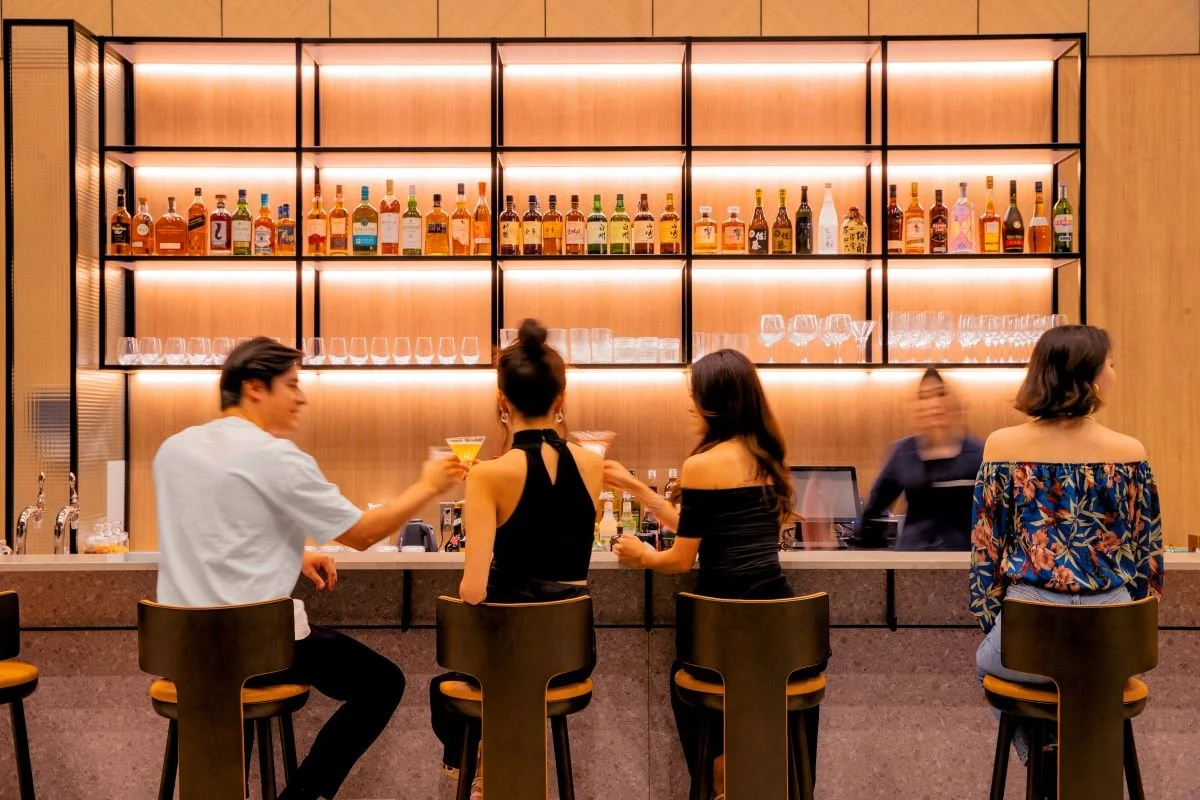 People sitting at a bar with a backlit alcohol shelf and barware behind them, engaging in conversation and drinks.