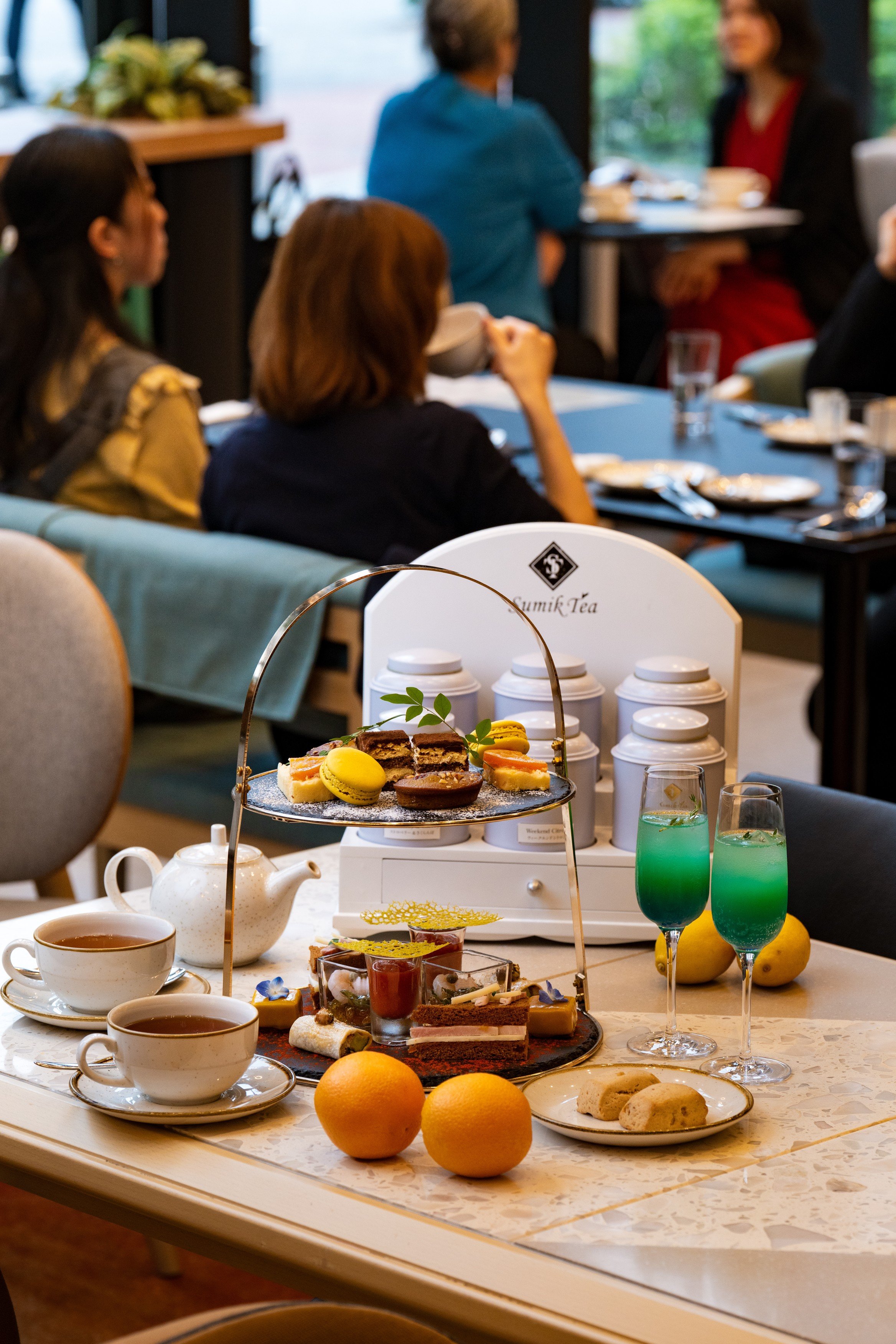 A three-tiered serving tray with assorted finger sandwiches and sweet treats, two glasses of iced tea with mint, a teapot with a cup of tea, and scones, all set on a dark table with a window in the background.