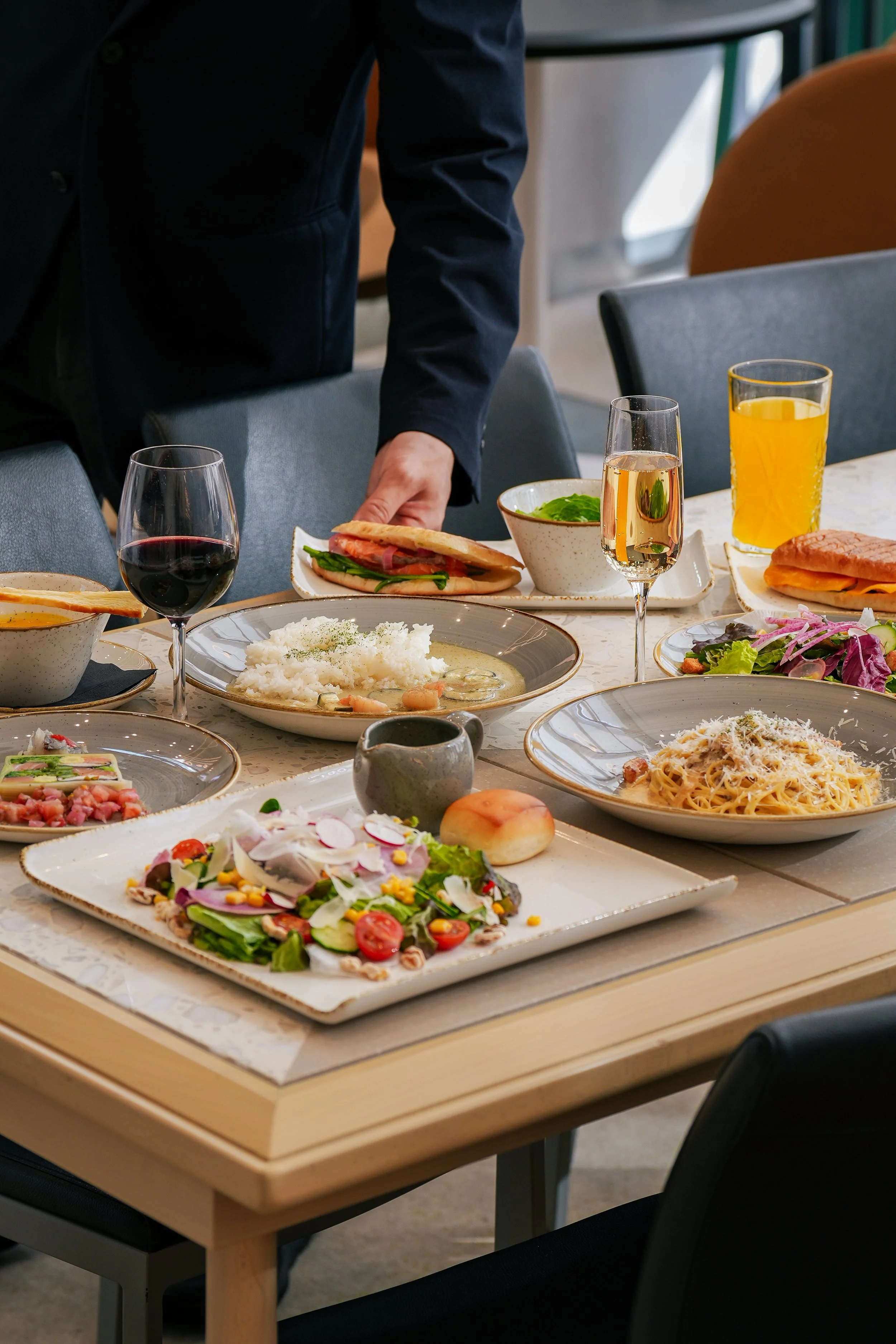 A person pouring salad dressing over a fresh vegetable salad that includes radish slices, cherry tomatoes, corn, cauliflower, green beans, and leaf garnishes, served on a white rectangular plate alongside a piece of toasted bread. In the background, there are bowls of soup, iced tea, a pasta dish, and another salad.