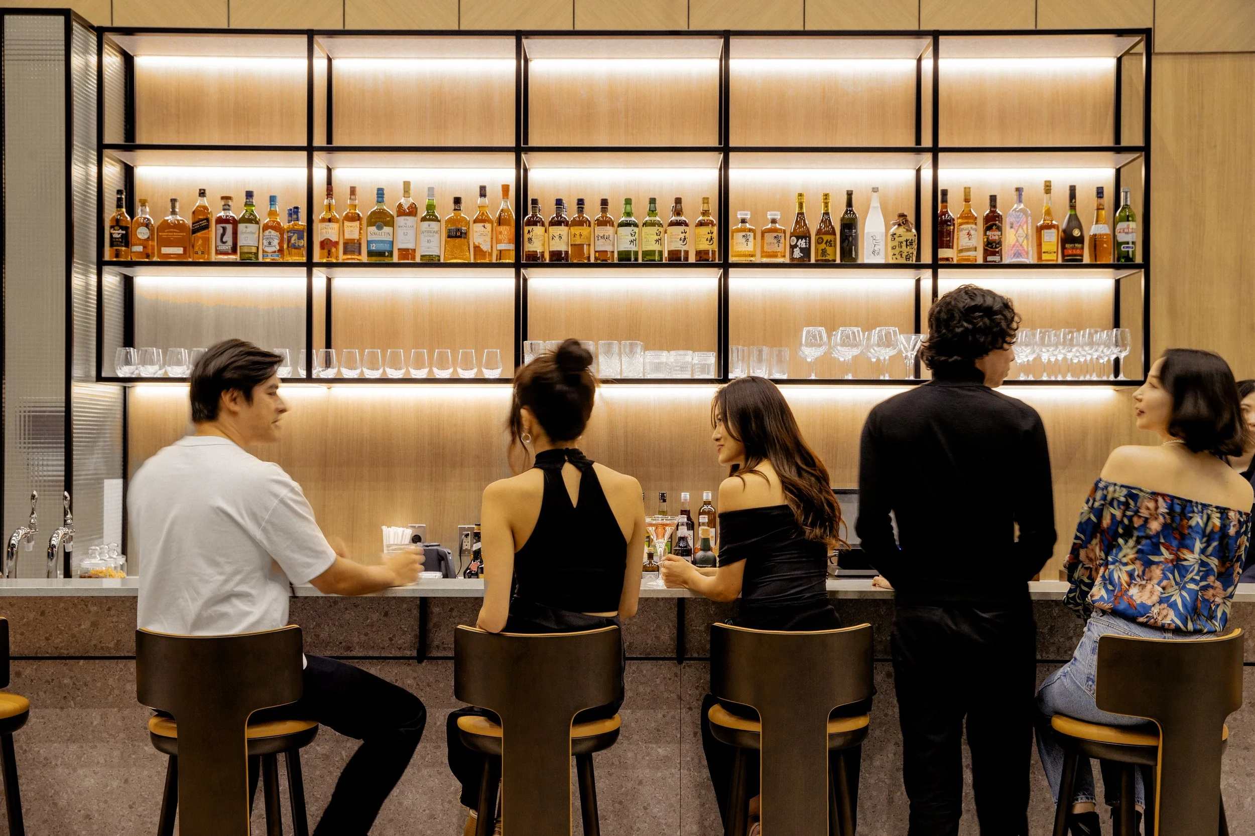 Stylish bar scene with five people sitting and standing at the counter, colorful liquor bottles, glasses, and a contemporary wooden and metal shelf backdrop.