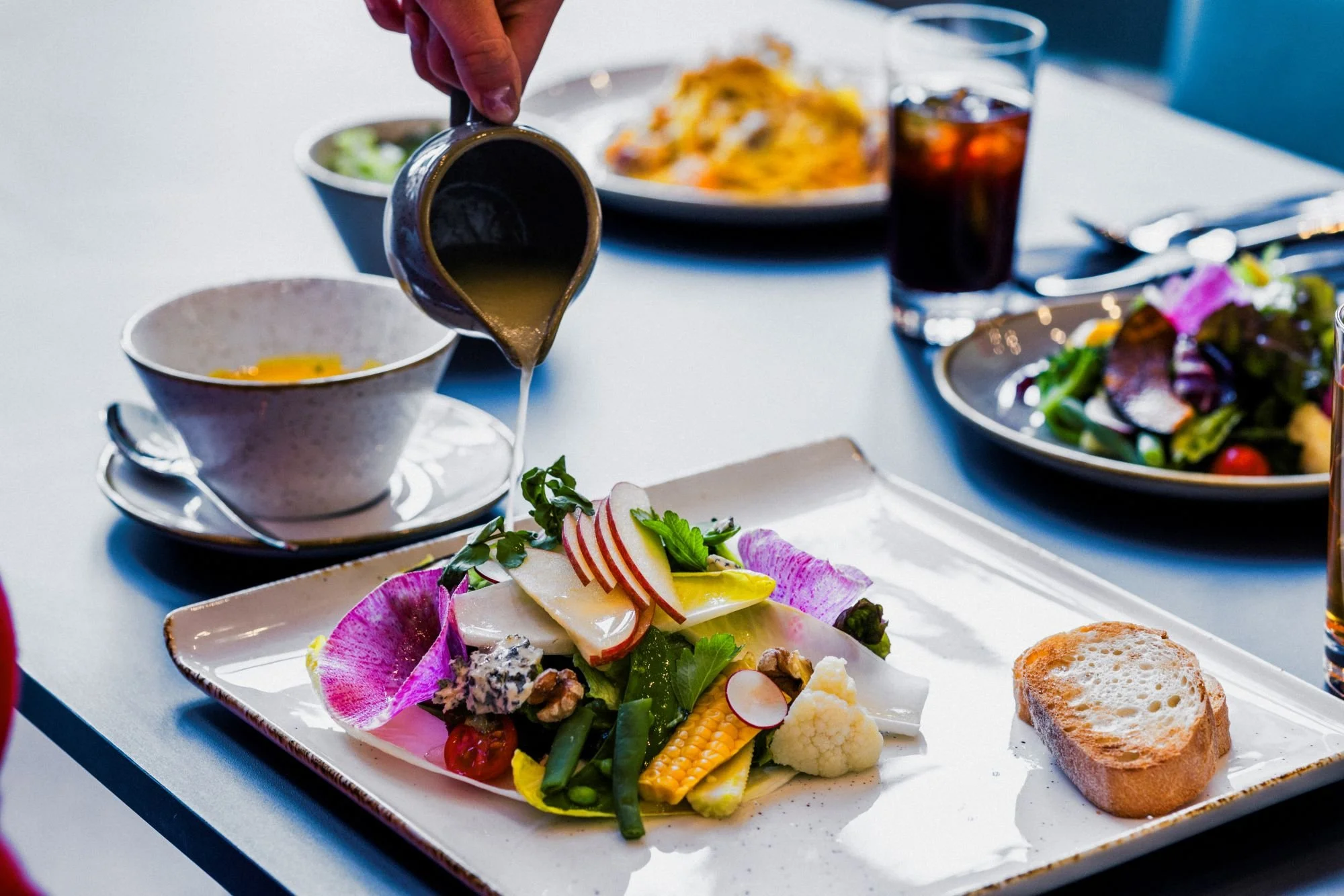 A person pouring salad dressing over a fresh vegetable salad that includes radish slices, cherry tomatoes, corn, cauliflower, green beans, and leaf garnishes, served on a white rectangular plate alongside a piece of toasted bread. In the background, there are bowls of soup, iced tea, a pasta dish, and another salad.