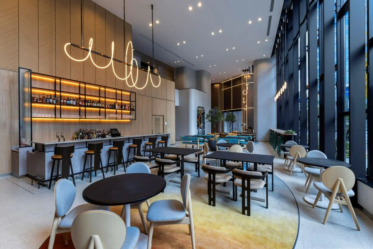 Modern bar and restaurant with high ceilings, large windows, and a mix of round and rectangular tables. A backlit bar with stools features liquor bottles, and there's stylish lighting and decor throughout, including potted plants and sleek furnishings.