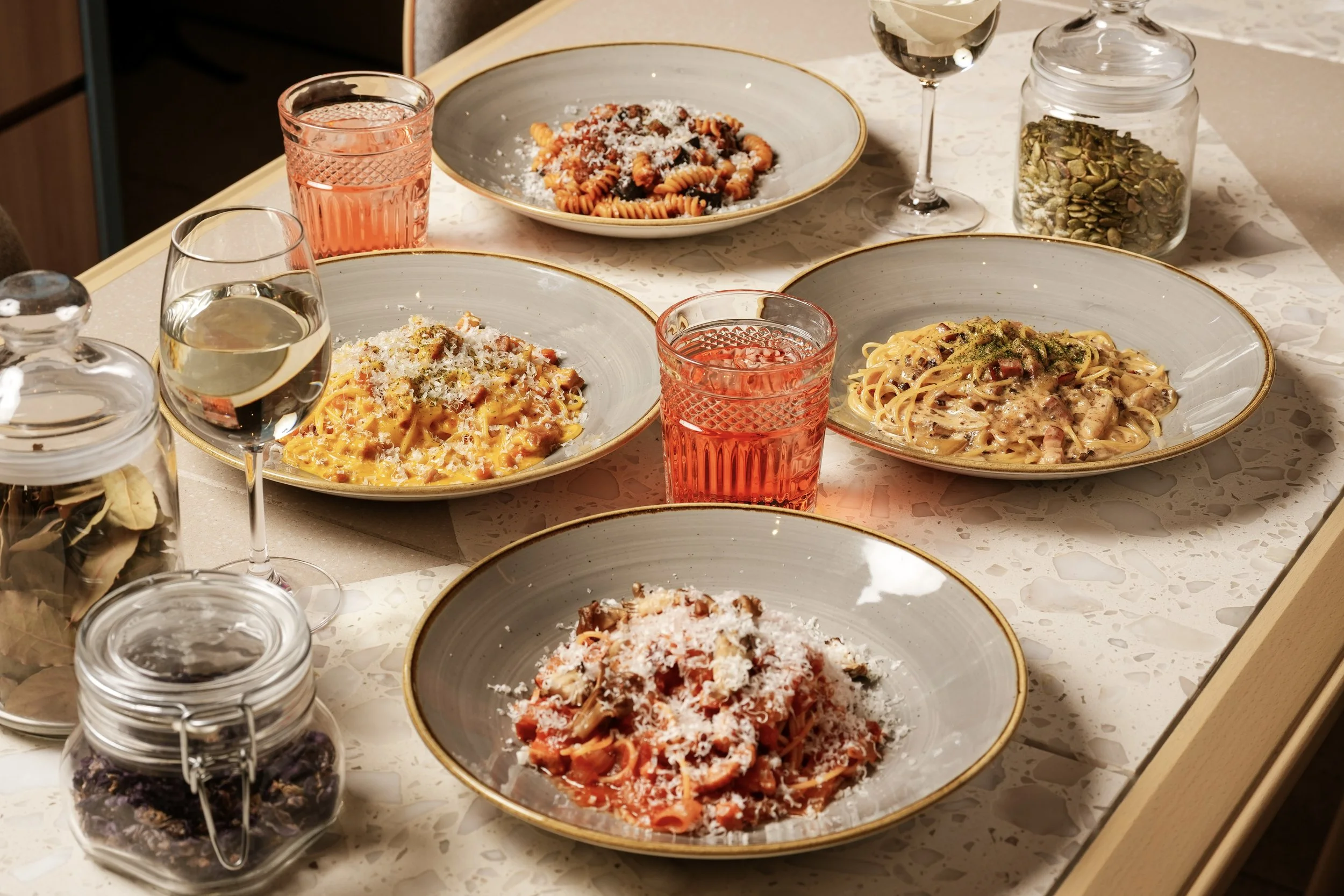 A plate of salad with shredded cheese, broccoli, ham, and tomatoes on a beige table with a glass of white wine in the background.