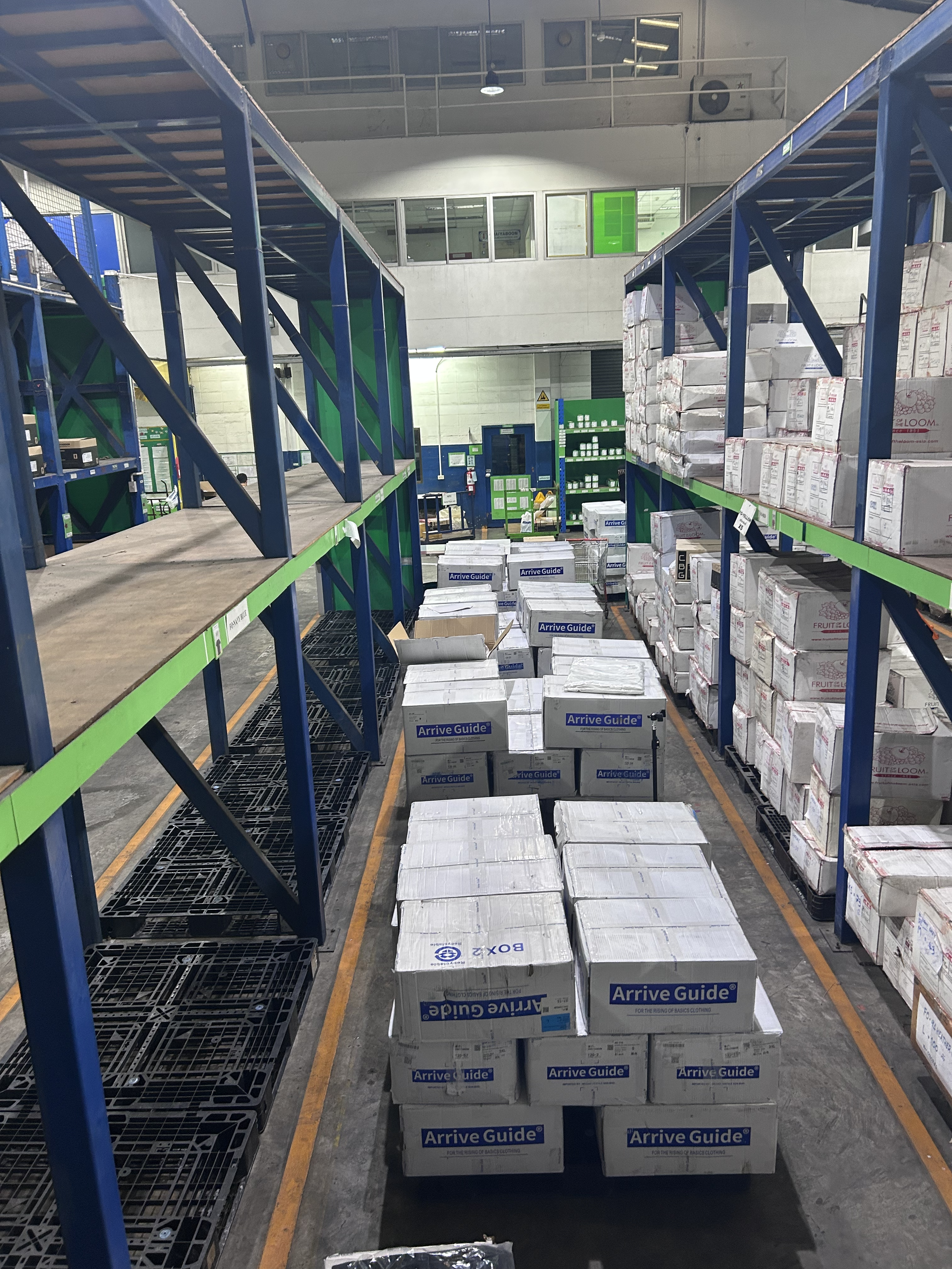Warehouse aisle with stacked boxes labeled 'Arrive Guide' and some on black pallets, metal shelving on either side, industrial lighting, and upper balcony area.
