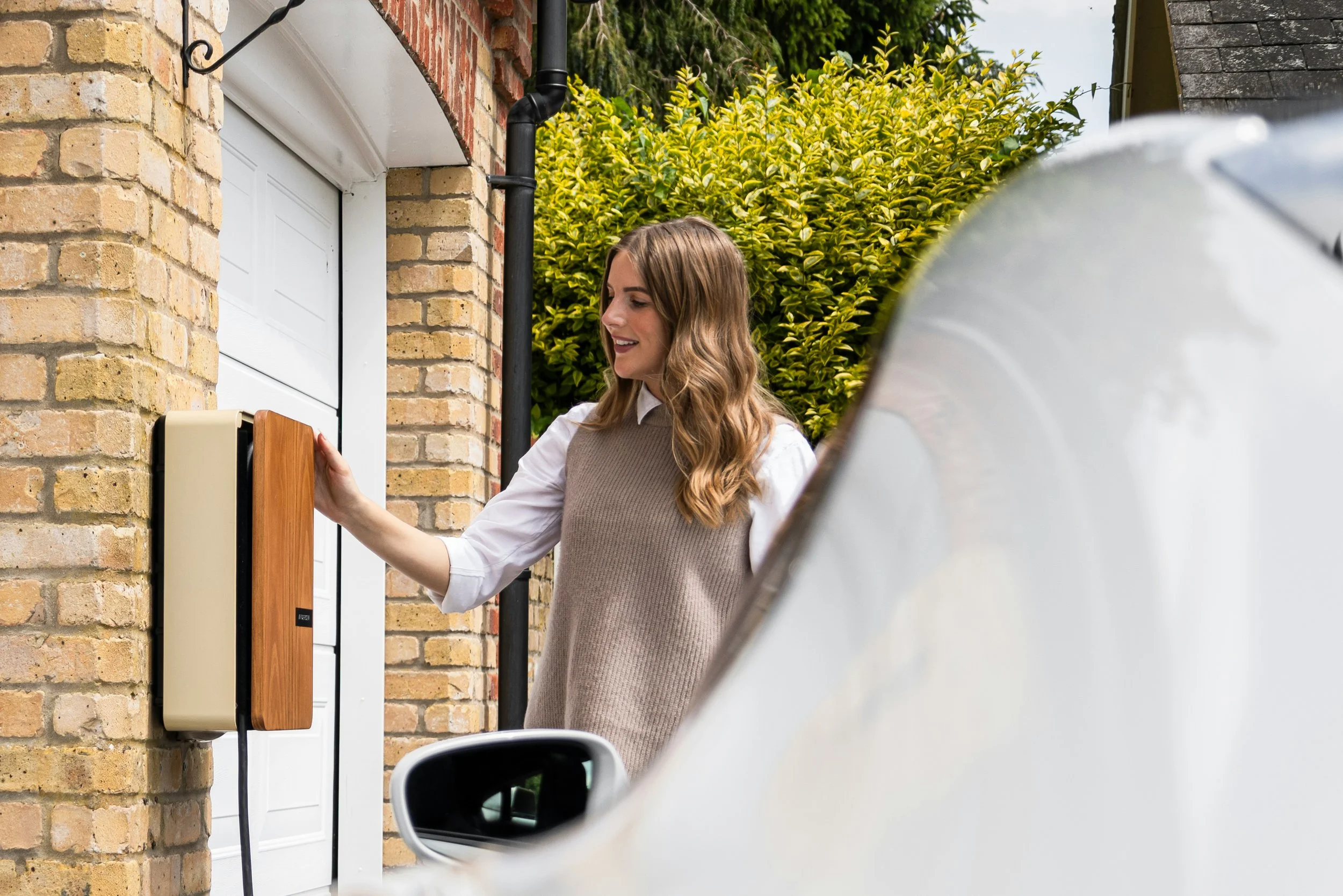 A woman with wavy brown hair hanging loose, wearing a white collared shirt and a beige sleeveless sweater, is reaching out to press a doorbell on a brick house. The house has a white door with an orange-brown wooden panel and a beige doorbell. The background shows a lush green hedge with yellow edges and neighboring rooftops. A white vehicle is partially visible in the foreground.
