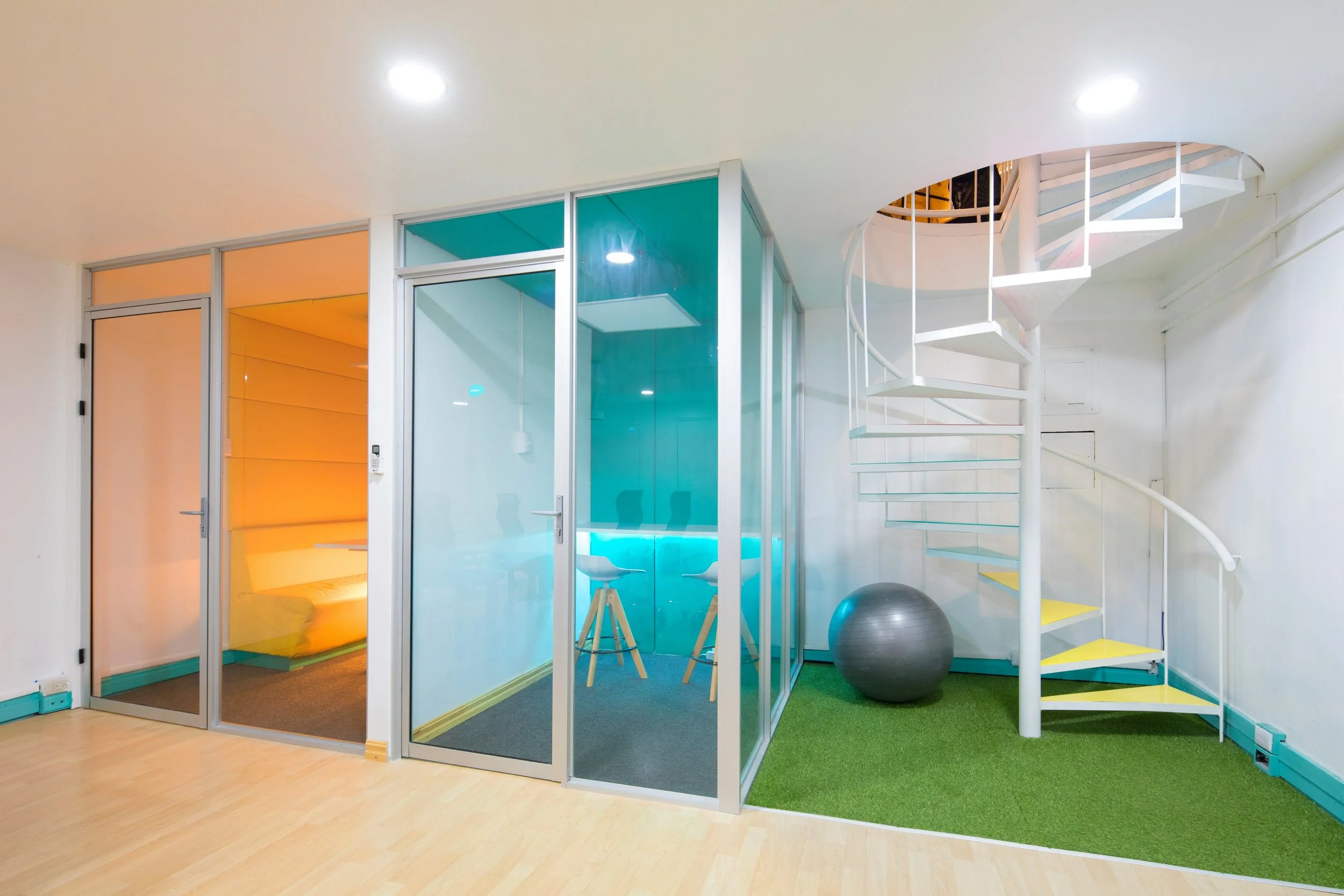 Interior view of a modern office or lounge area with a spiral staircase, glass enclosed meeting rooms with colorful walls, a black exercise ball, and green carpeted flooring.